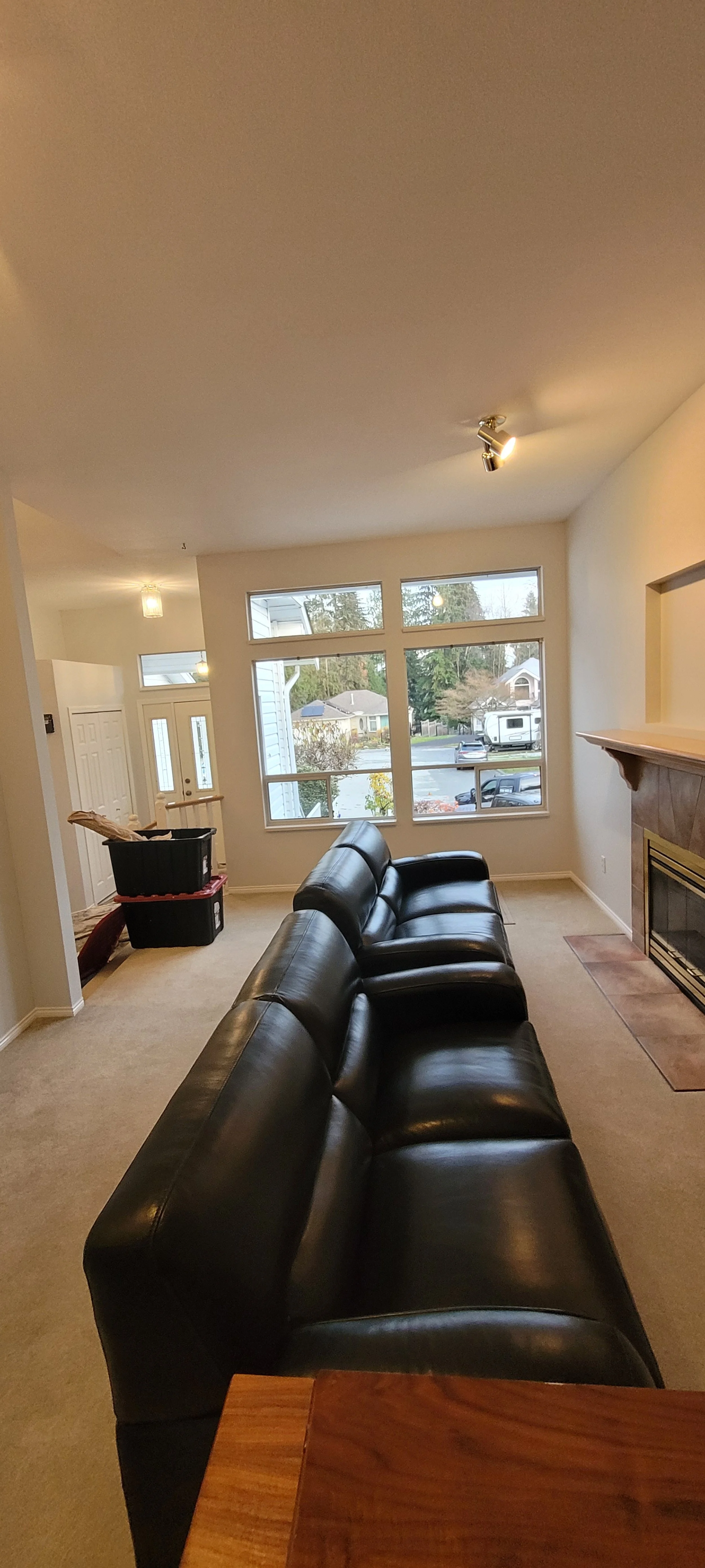 Living room with black leather couch, large window overlooking suburban neighborhood, fireplace, and box with construction materials.