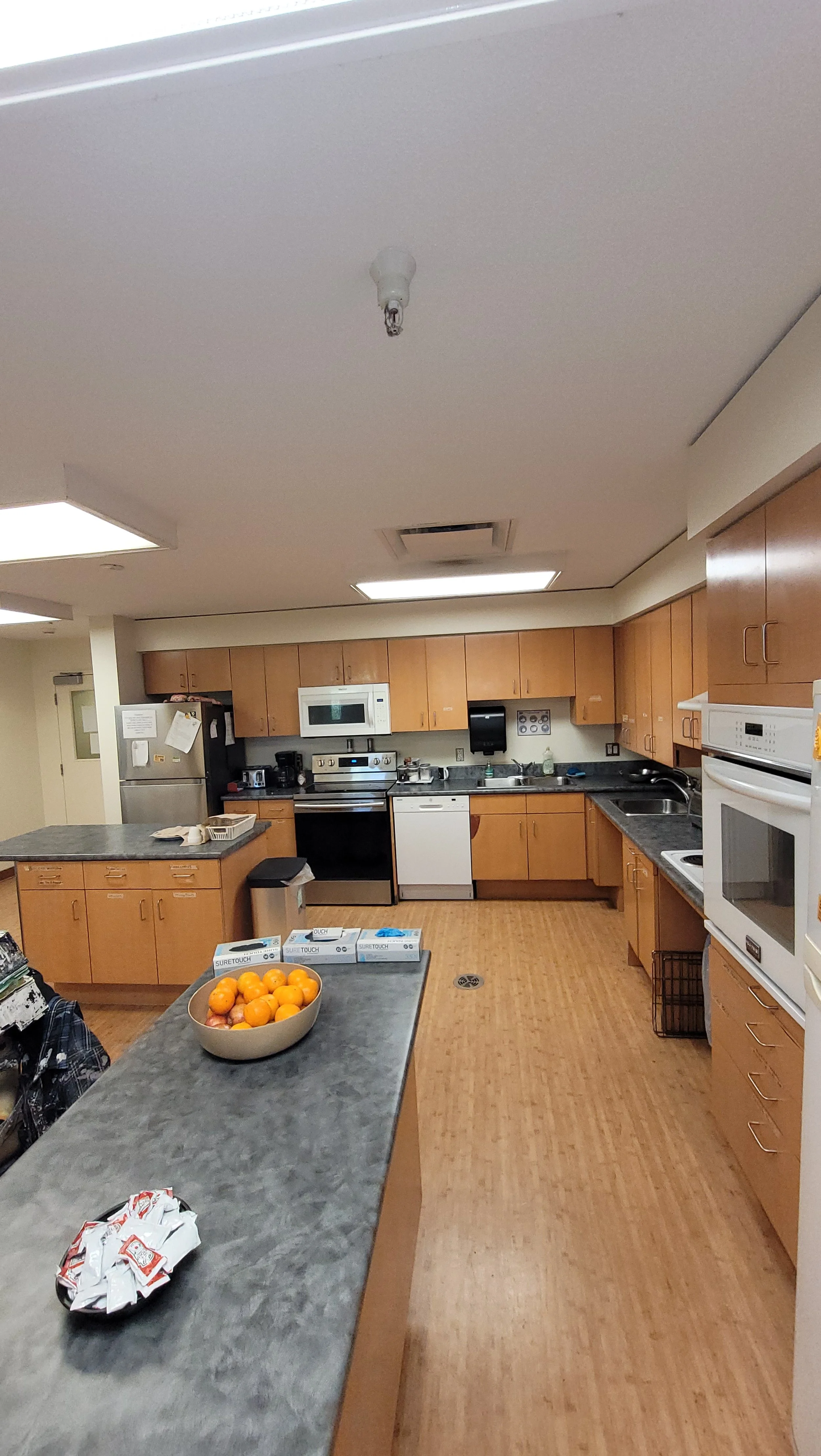 Kitchen with wooden cabinets, white appliances, and a gray countertop. Bowl of oranges on the island, various small appliances and papers.