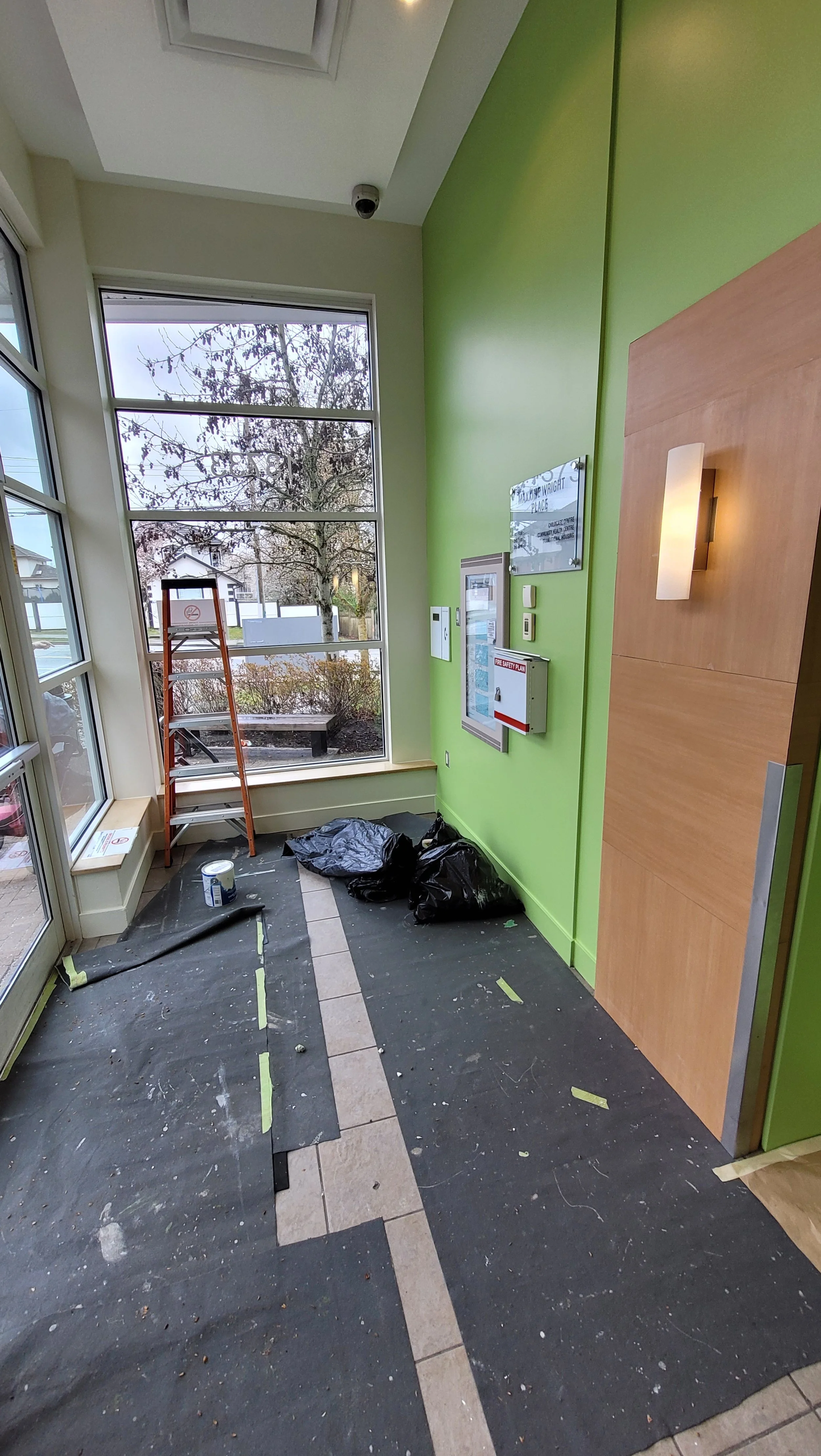Interior of a building with a green wall, large window, ladder, and cleaning supplies on the floor, indicating ongoing cleaning or maintenance work.