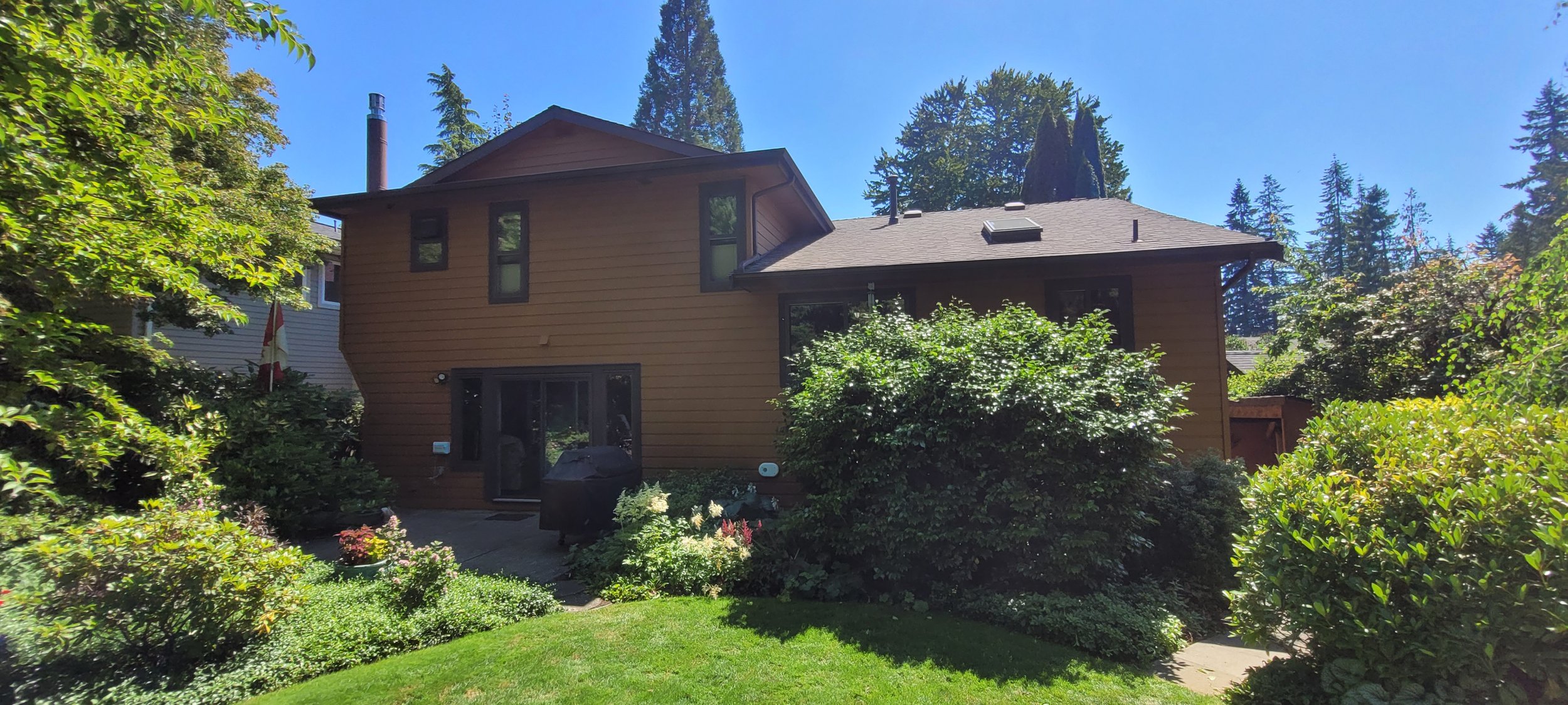 Backyard view of a two-story tan house with several windows, surrounded by lush green bushes, trees, and a well-maintained lawn, under a bright blue sky.