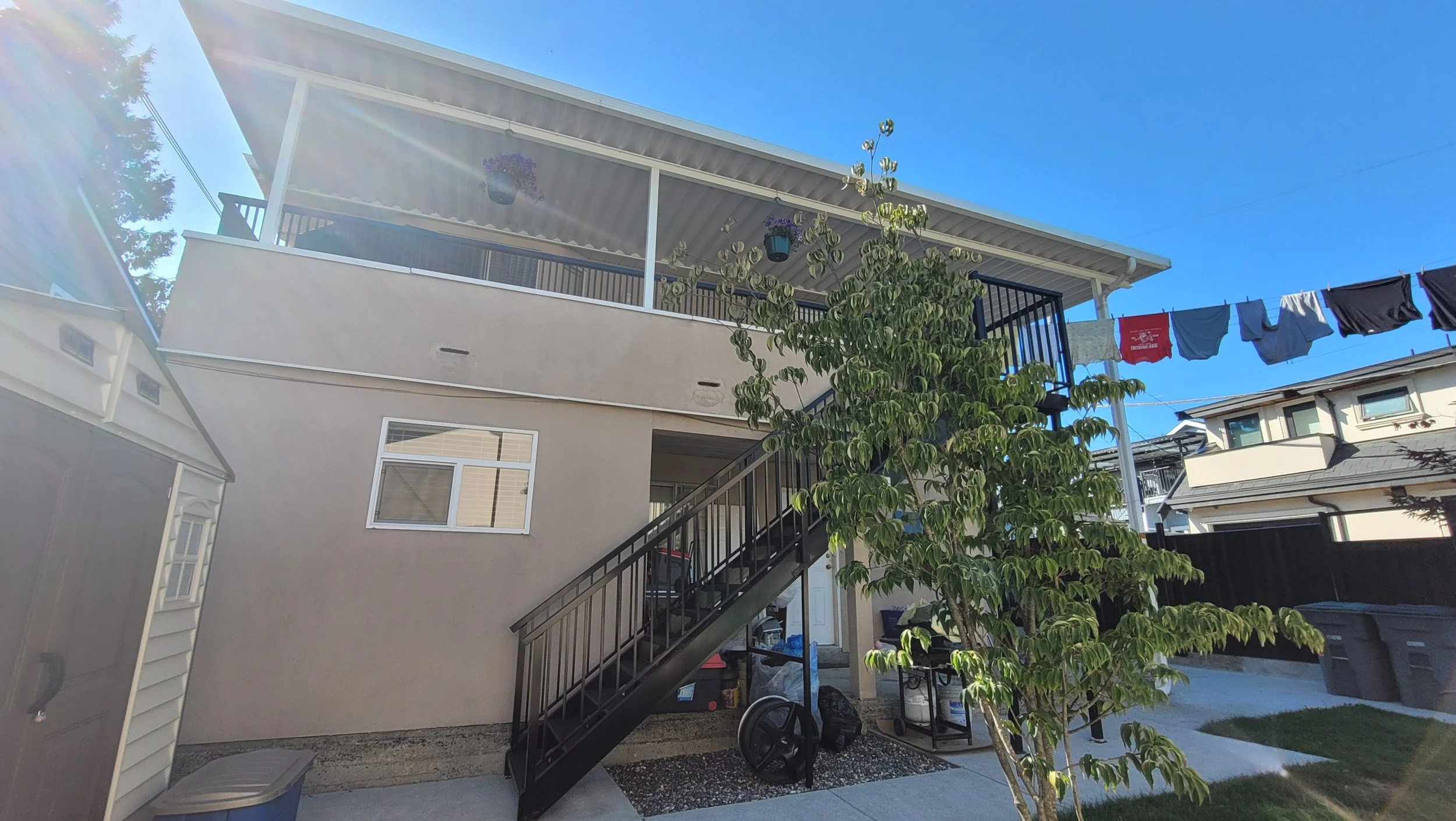 Backyard with stairs leading to an upper balcony, laundry hanging on a clothesline, and a small tree in the foreground under a clear blue sky.