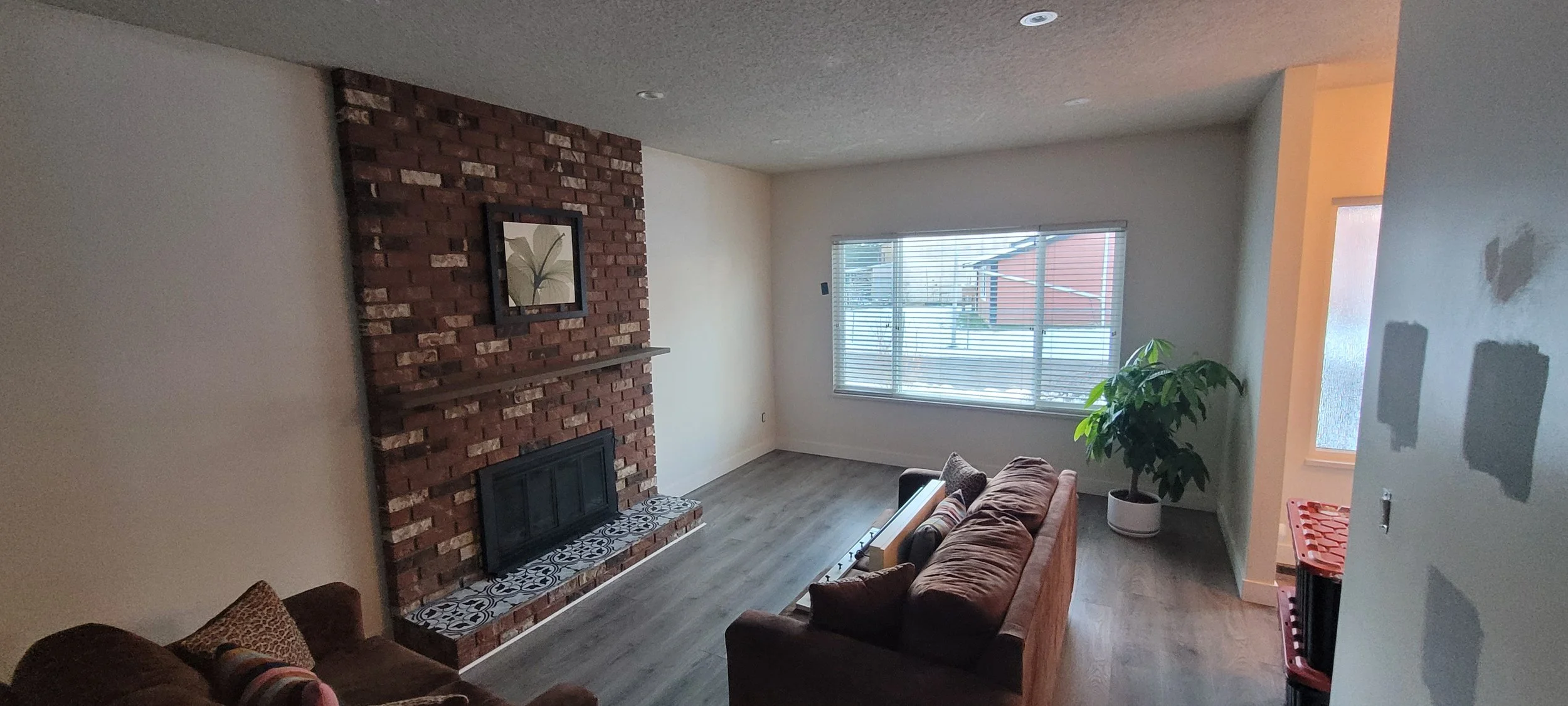 Living room with a brick fireplace, a window with blinds, a brown couch, a potted plant, and some small furniture near the wall.