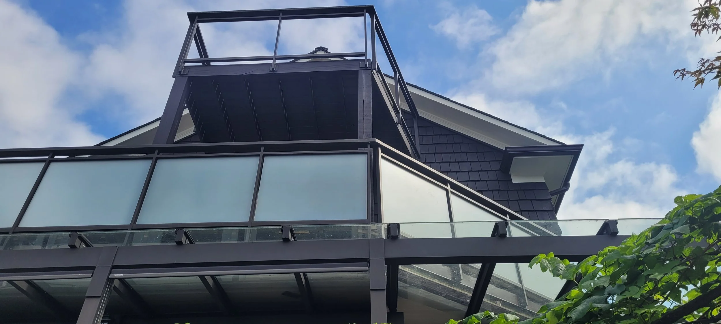 Upward view of a modern building with glass balconies and dark exterior siding, against a partly cloudy sky, with green foliage in the foreground.