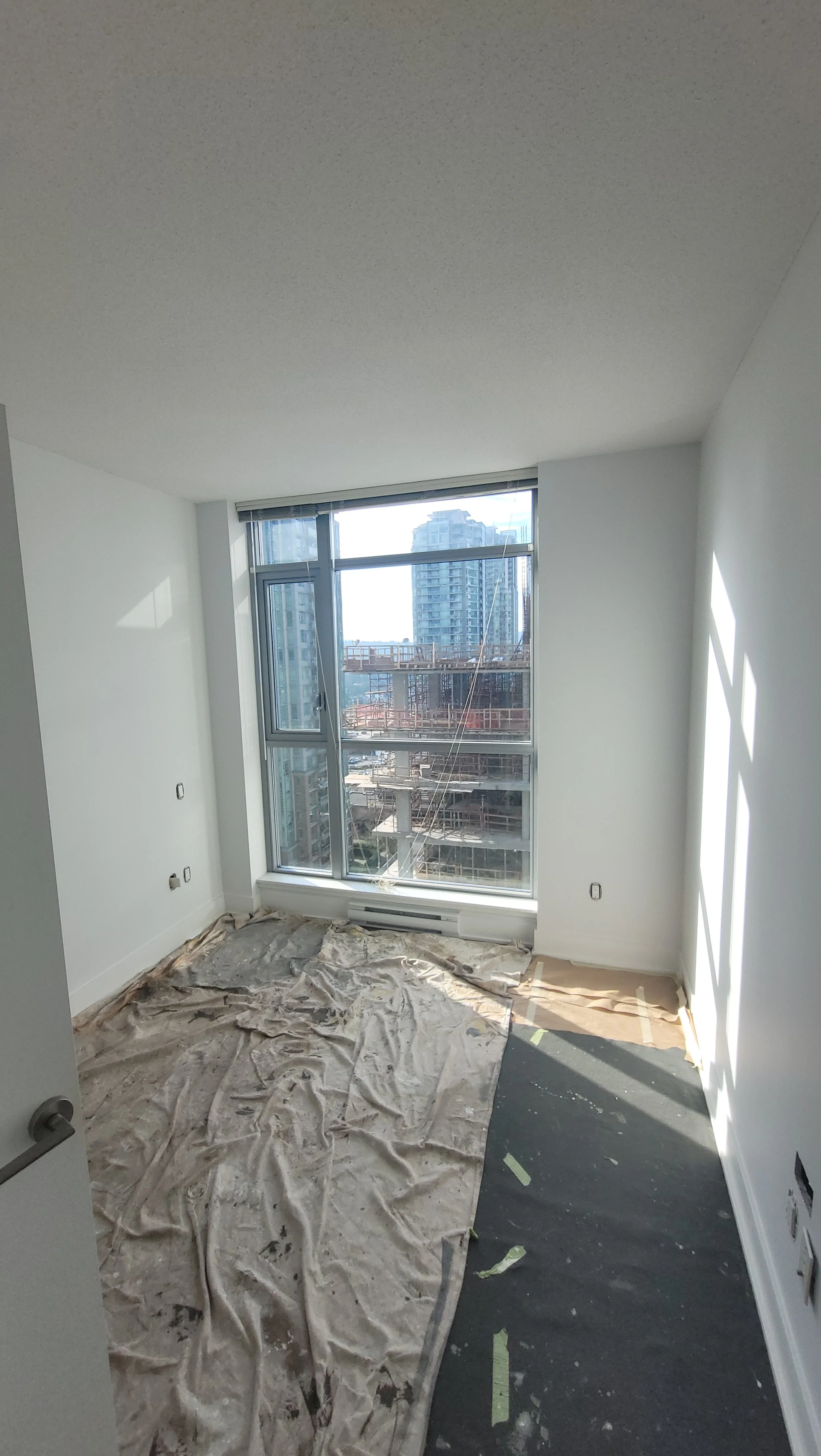 Empty room with construction materials, partially covered with a tarp, large window showing city buildings and a construction site outside.