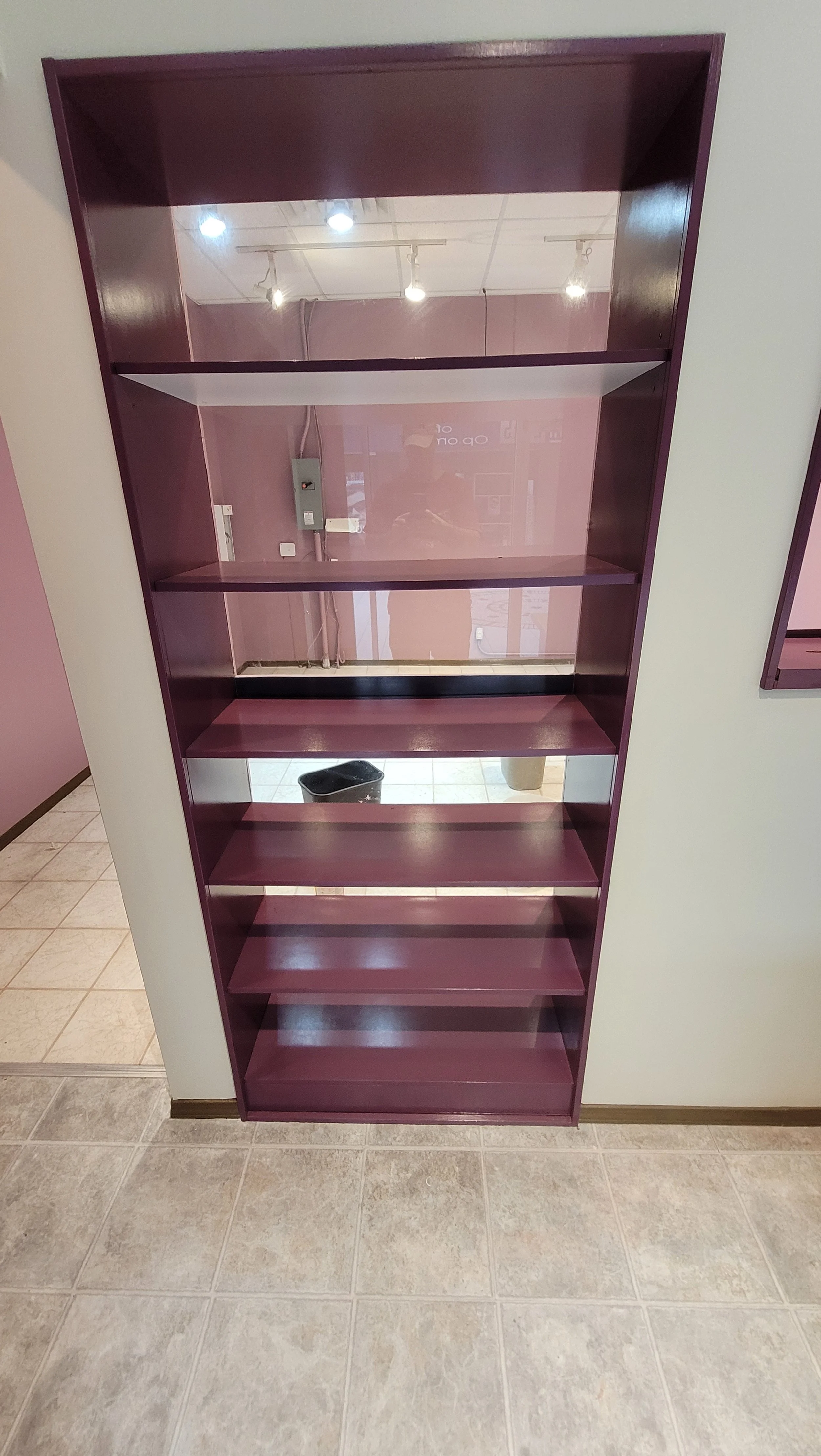 Empty purple bookshelf with multiple shelves, located indoors near a beige wall and tiled floor, with a mirror reflecting part of the room.