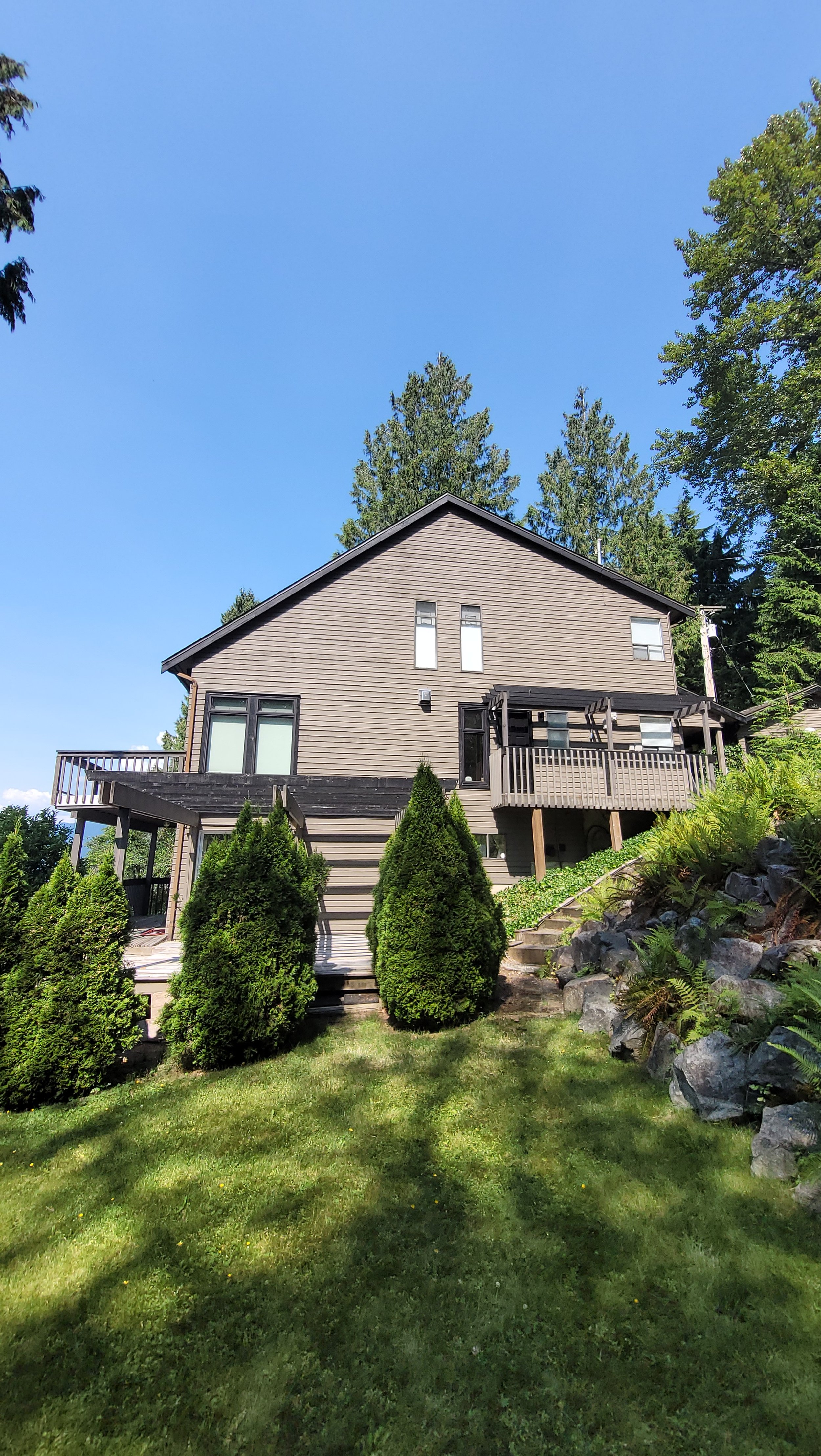 A house with a steep roof, brown wooden siding, and multiple windows, surrounded by green bushes, trees, and a grassy yard under a clear blue sky.