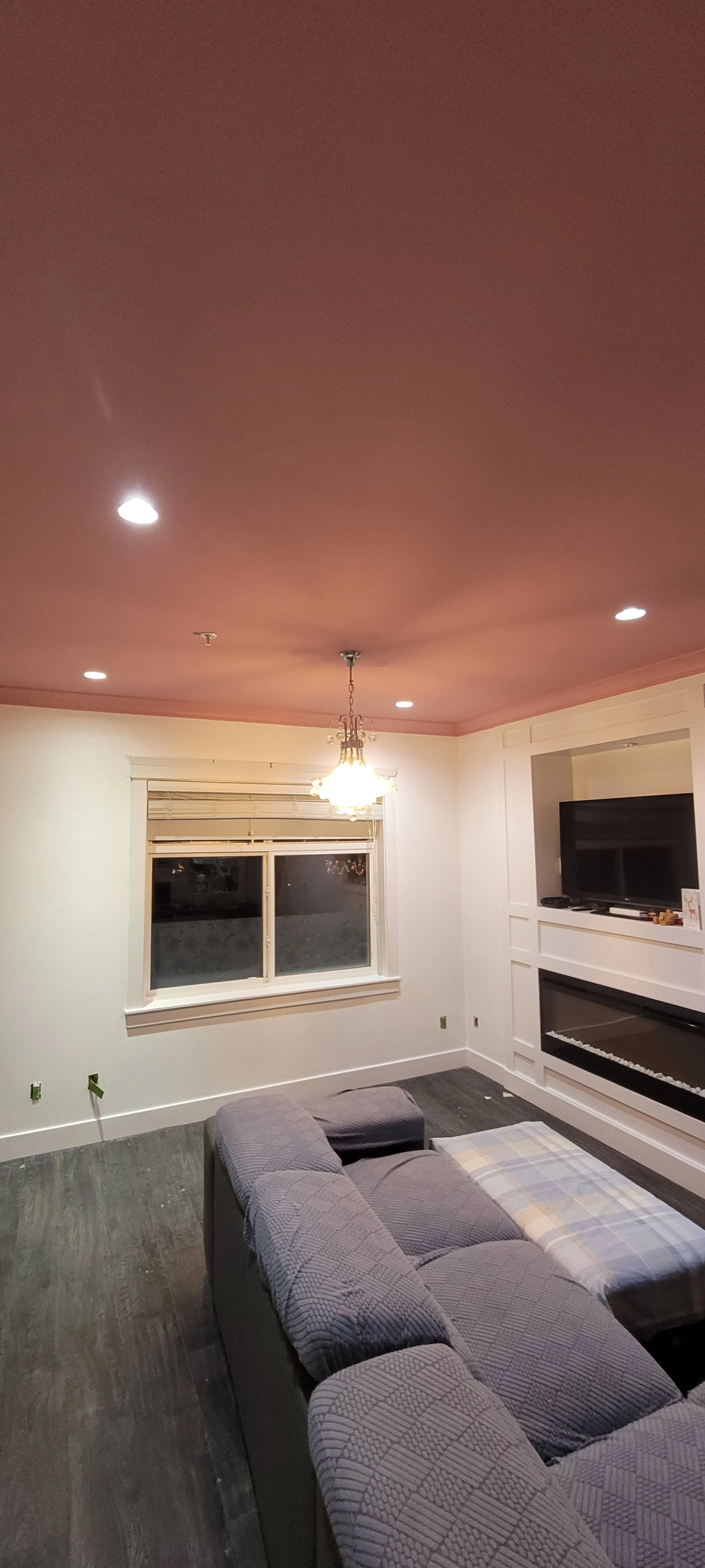Living room with a pink ceiling, white walls, a window with blinds, a chandelier hanging from the ceiling, a flat-screen TV on a white entertainment unit, and a gray sectional sofa.