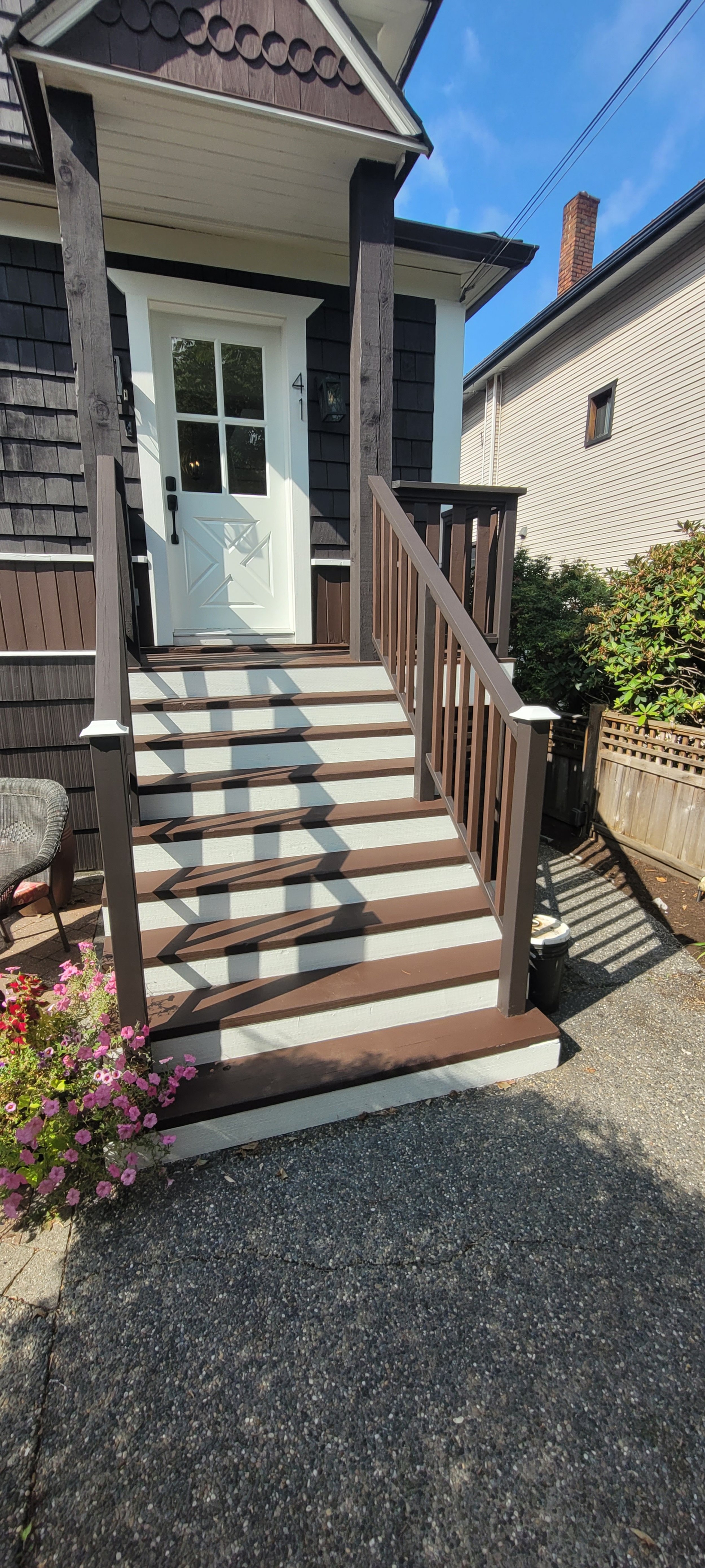 Front porch with steps leading to a white door, black shingle siding, and a small porch roof. There are shadows on the steps and flowers on the left side.