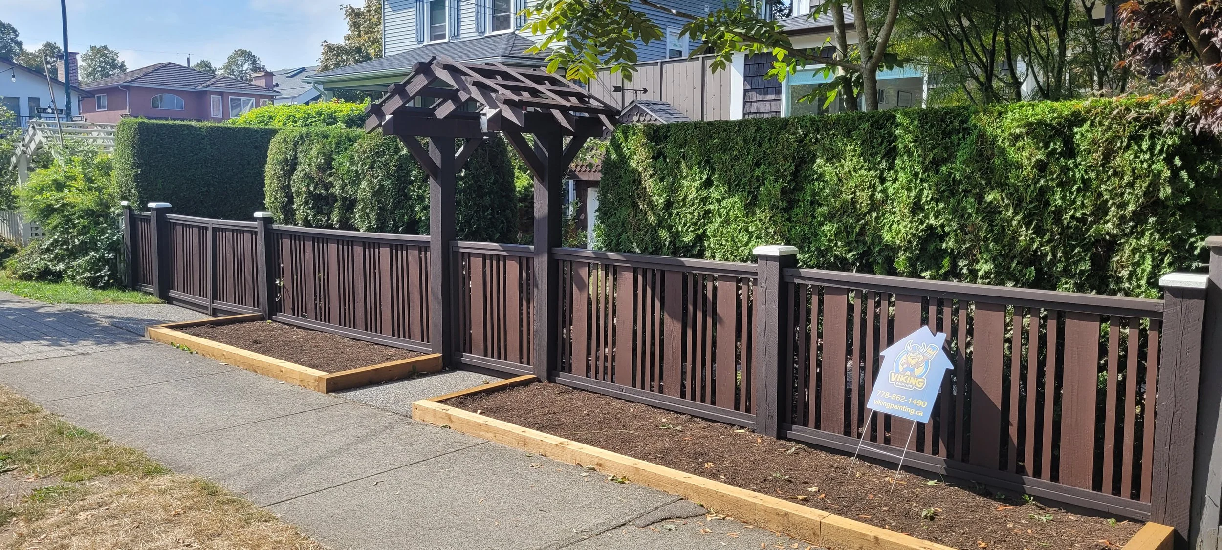 A backyard with a newly installed wooden fence and two garden beds bordered with wood, located on a sidewalk. There is a sign in the right garden bed with a Viking logo and contact information.