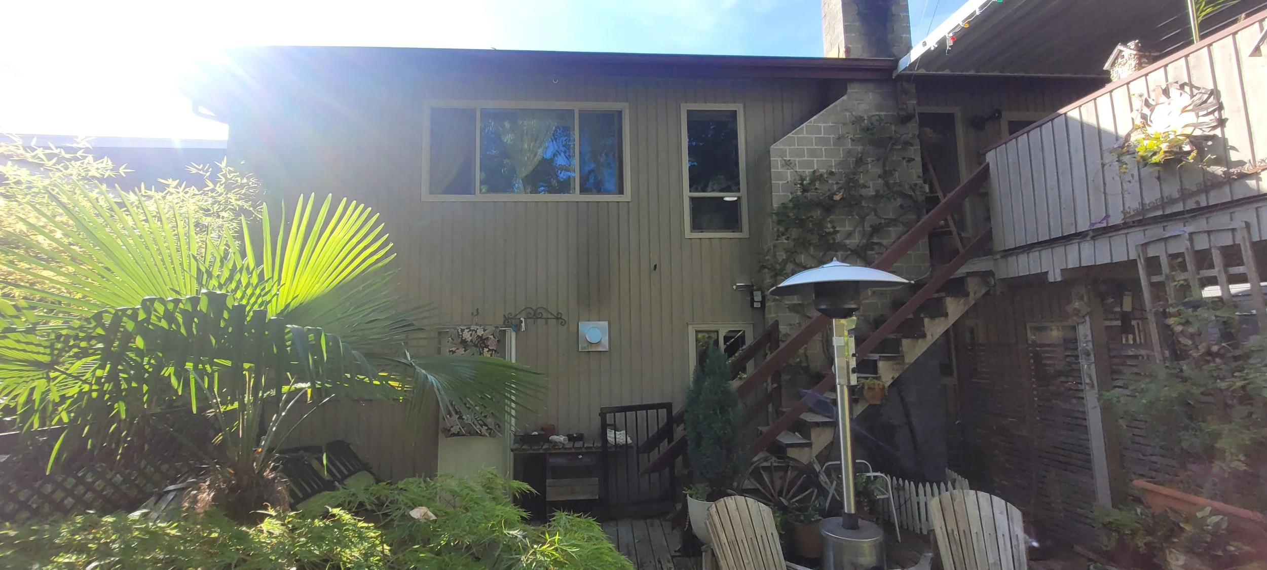 Backyard patio with outdoor furniture, large palm plant, patio heater, stairs leading to an upper deck, and a beige house with windows.
