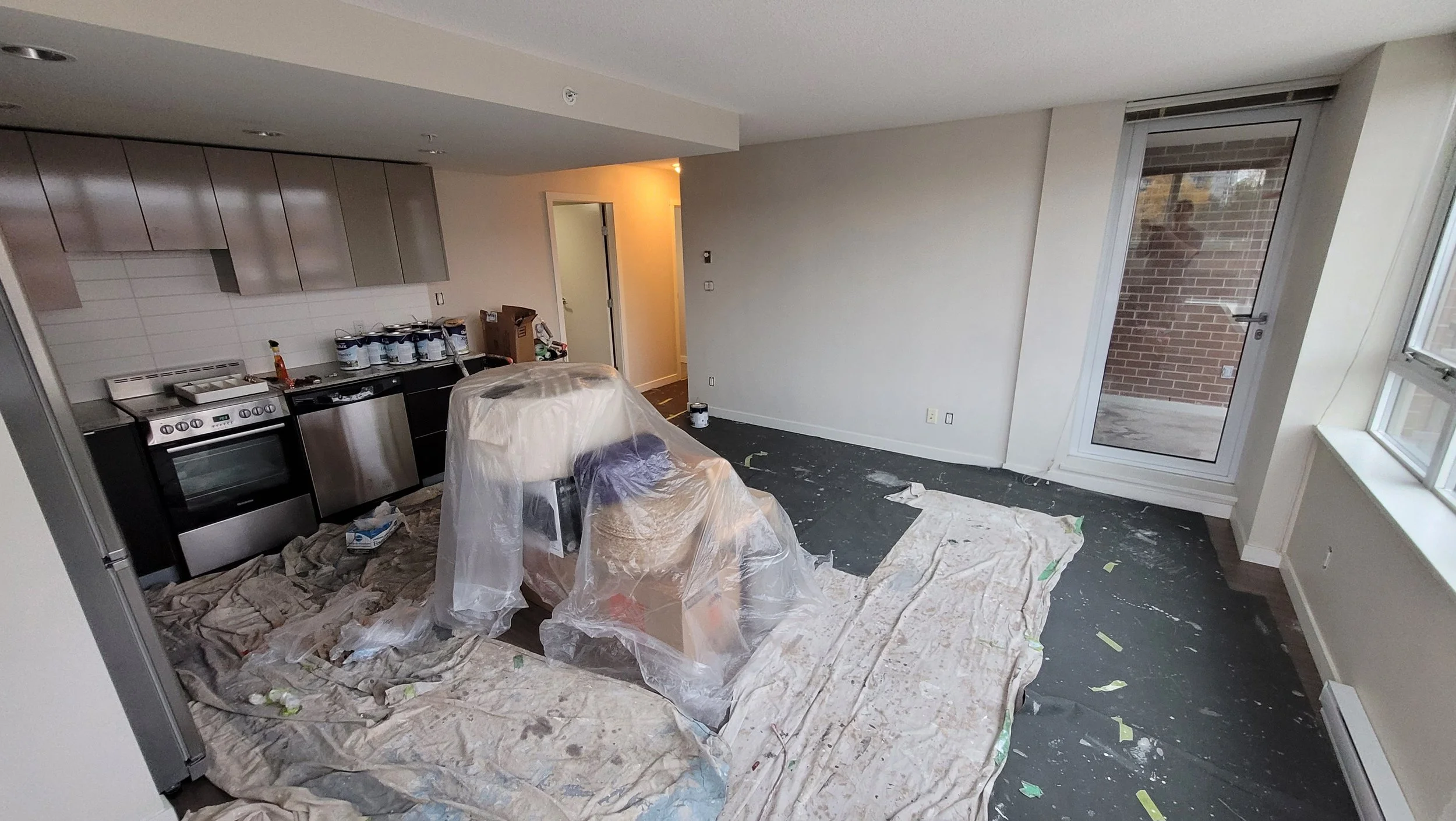 Room under renovation with plastic covering furniture, paint cans lined up along the kitchen counter, and painting supplies on the floor and window sills.