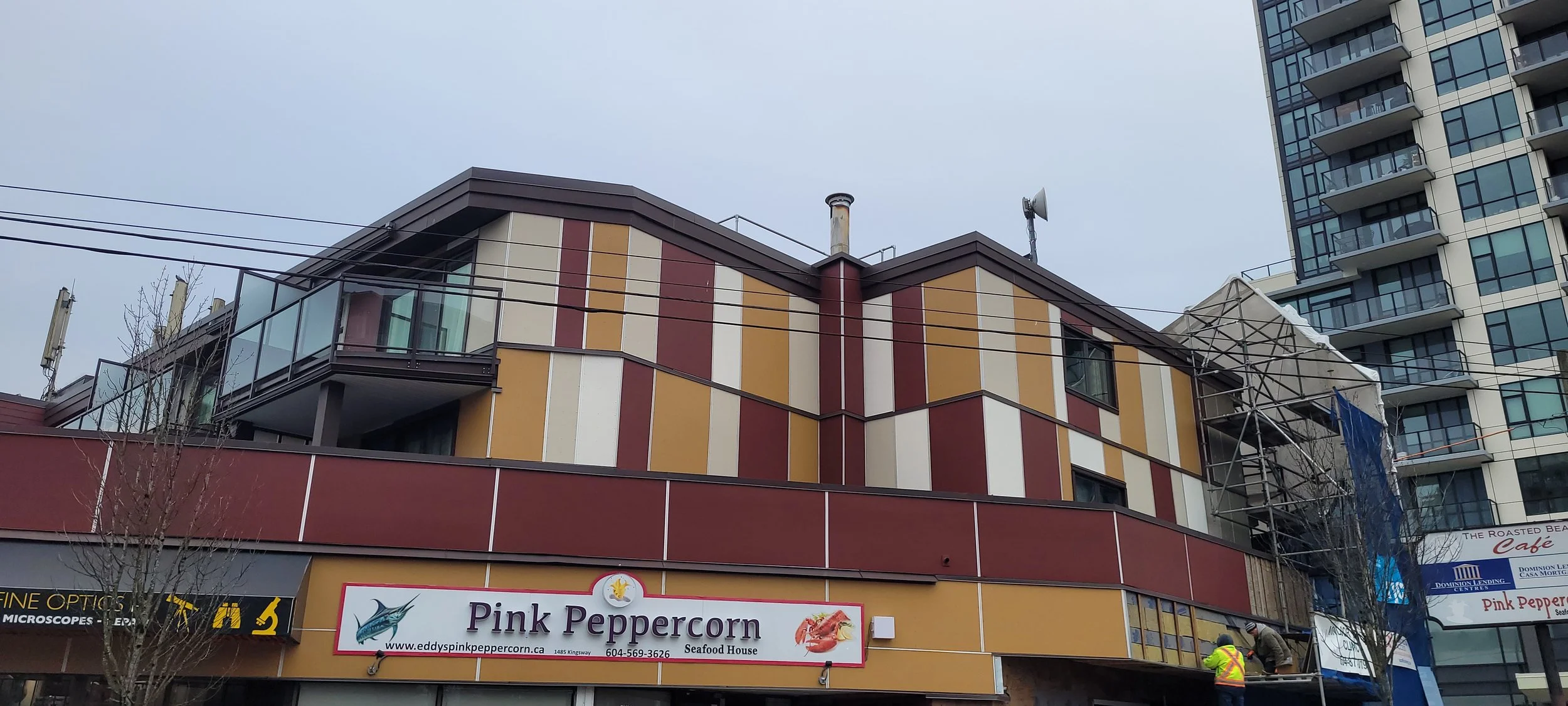 Modern multi-story building with colorful paneling and a seafood restaurant sign for Pink Peppercorn. Construction scaffolding on the right side, workers in safety vests, and neighboring high-rise buildings.