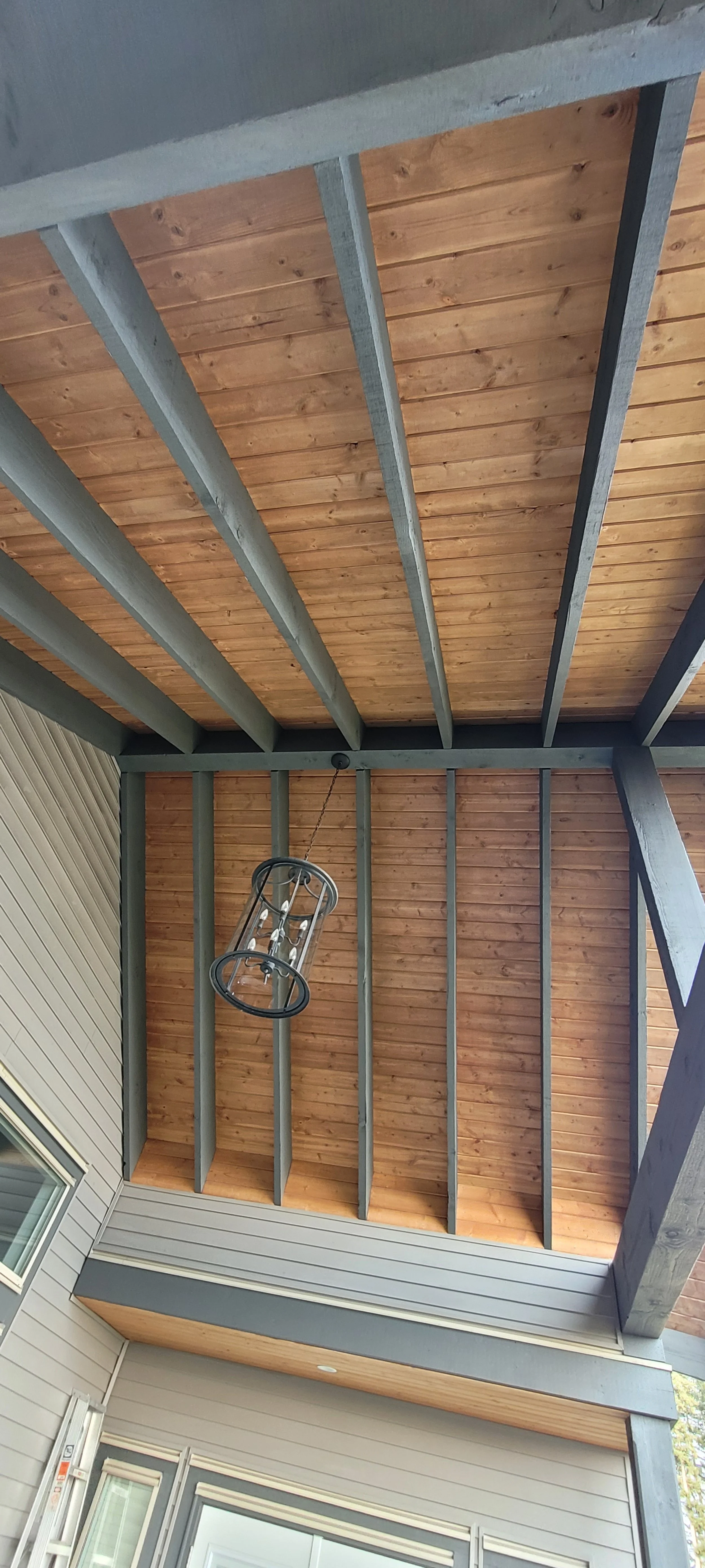 View of a covered porch ceiling with wooden planks and exposed beams, featuring a hanging lantern-style light fixture.