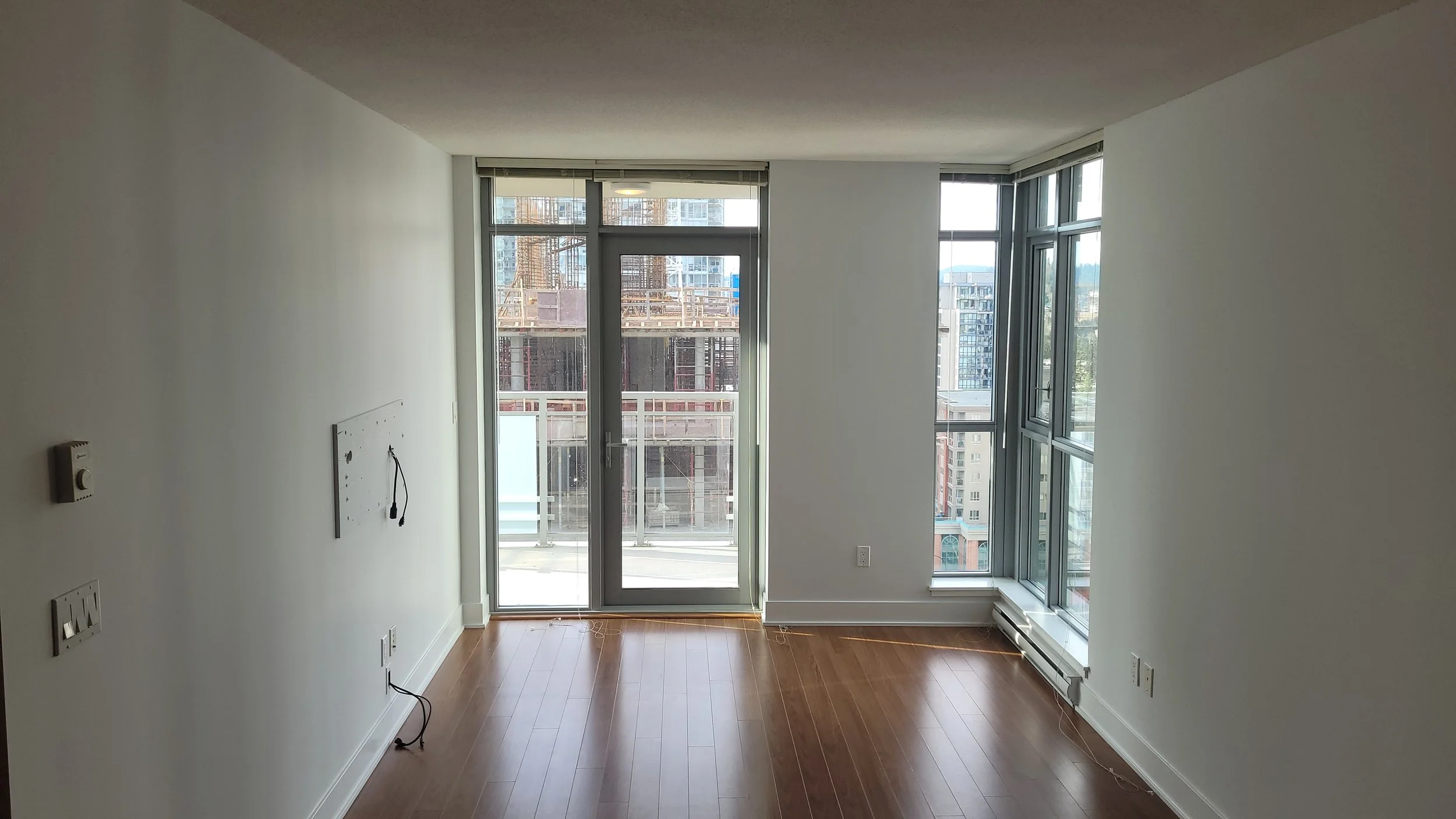 Empty apartment room with large glass windows and a door leading to a balcony, wooden flooring, and a cityscape view outside.