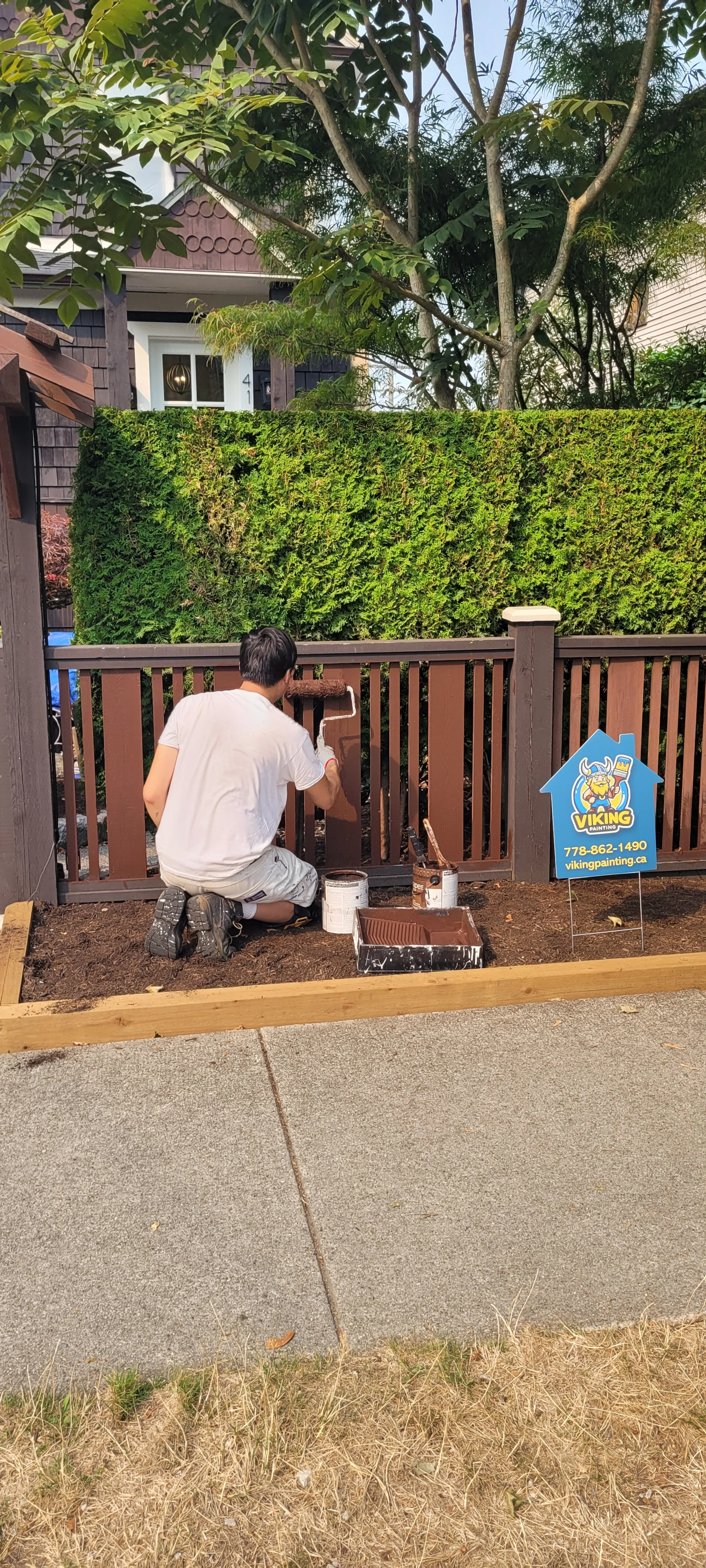 A person kneeling on the ground painting a wooden fence with brown paint, with painting tools nearby, in front of a green hedge, during daytime.