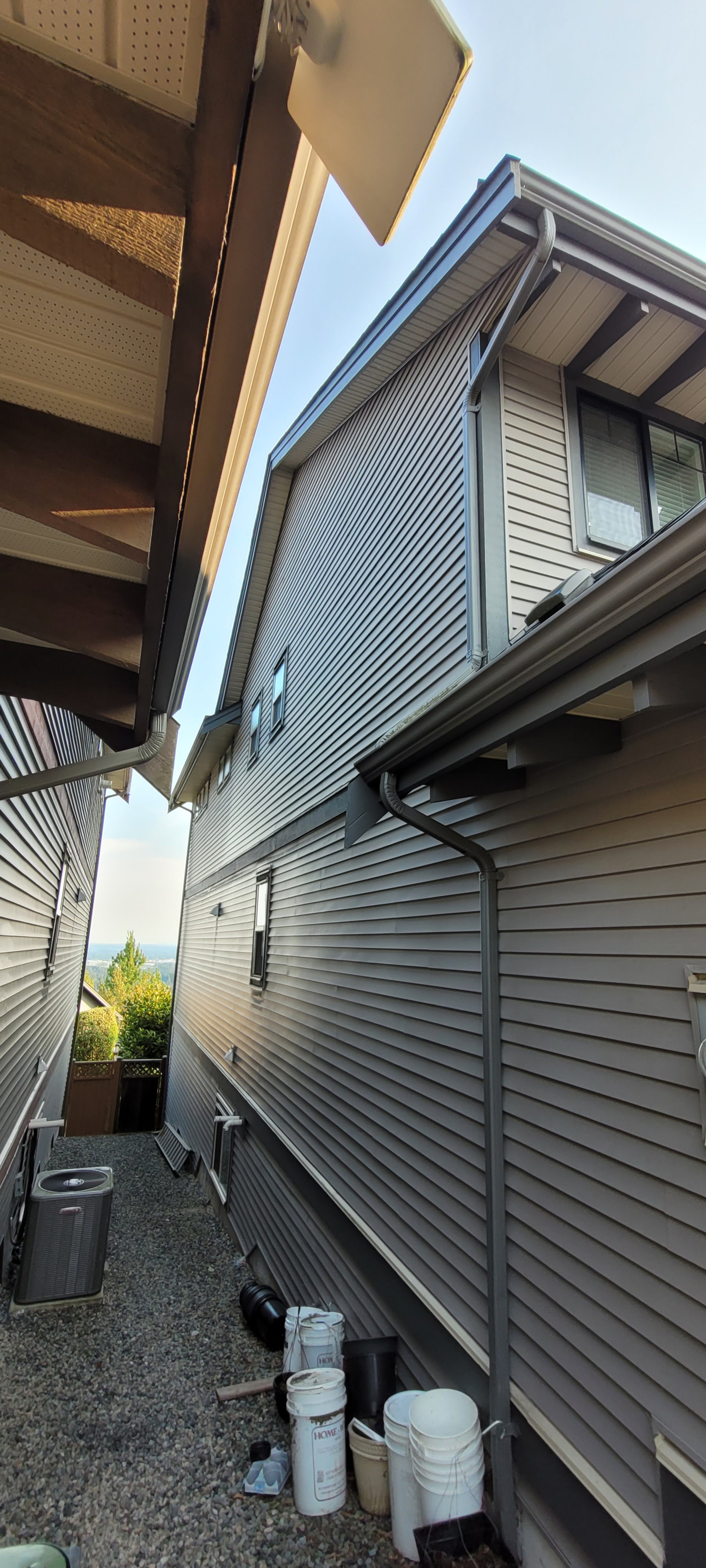 An alleyway between two beige vinyl-sided houses, with visible gutters, downspouts, and a gravel-covered ground. Buckets and a few other objects are on the ground, and there is an HVAC unit on the left side.