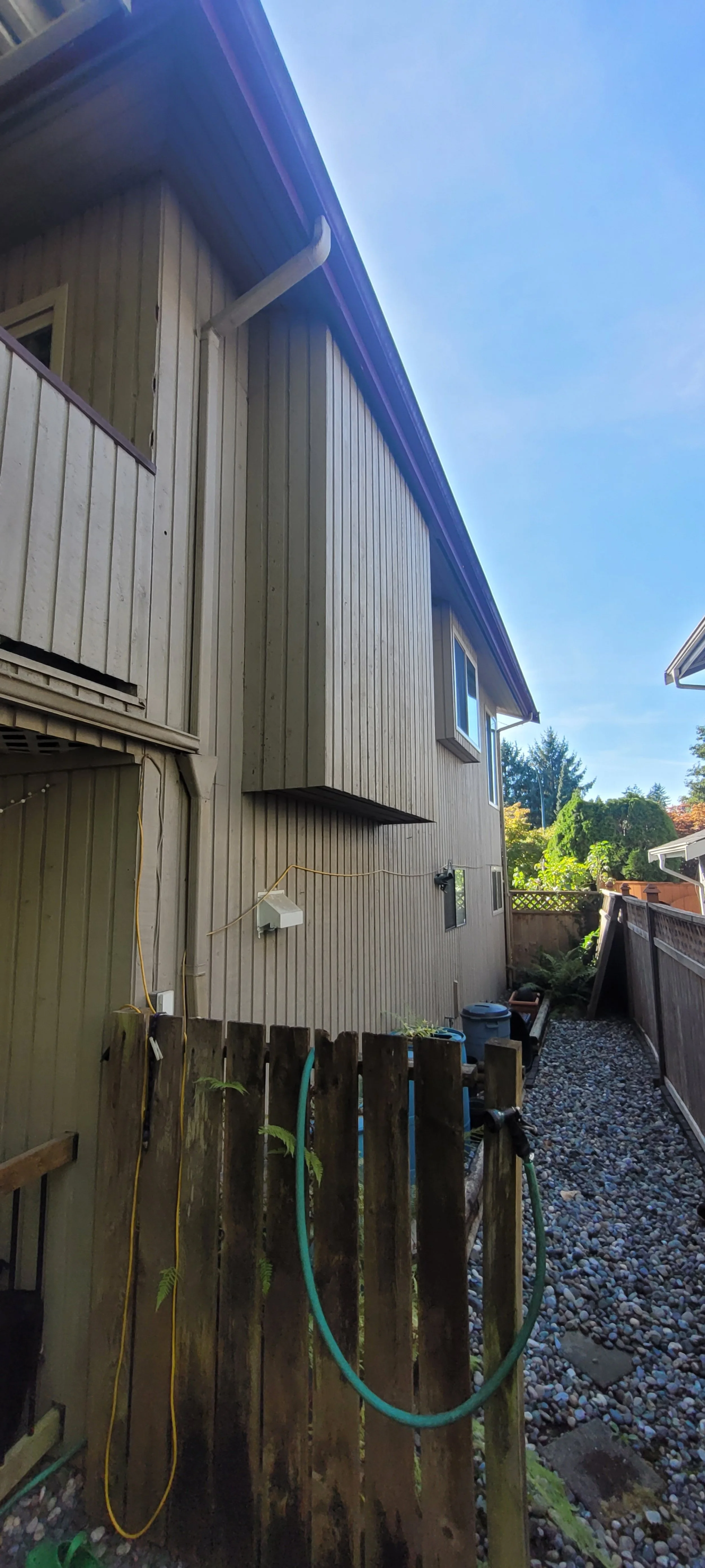 View of the side of a beige house with vertical siding, small windows, and a dark-colored roof. There is a narrow gravel path along the house, enclosed by a wooden fence, with gardening tools and water hoses visible.