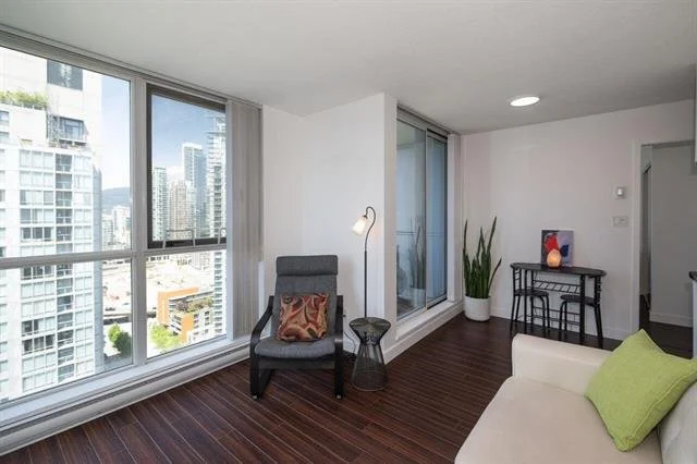 Modern living room with large windows overlooking a city skyline, dark hardwood floors, gray armchair with a colorful pillow, floor lamp, tall plant, small desk with chair, white sofa with green pillow, and an abstract painting.