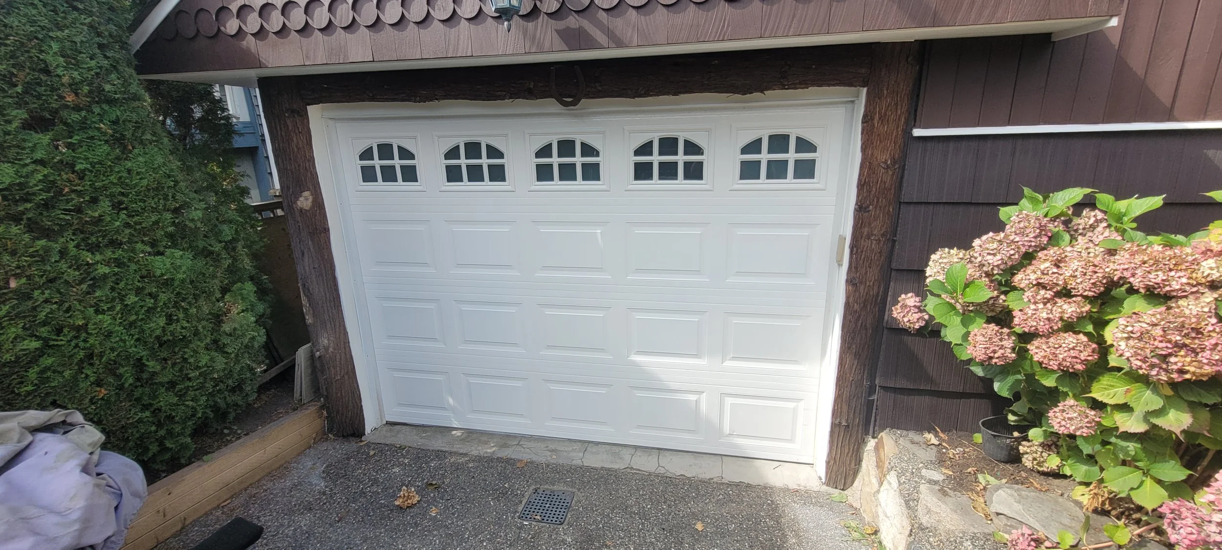 A white garage door with four small windows at the top, surrounded by brown wood siding, a lush green hedge to the left, and pink hydrangea flowers with green leaves to the right.