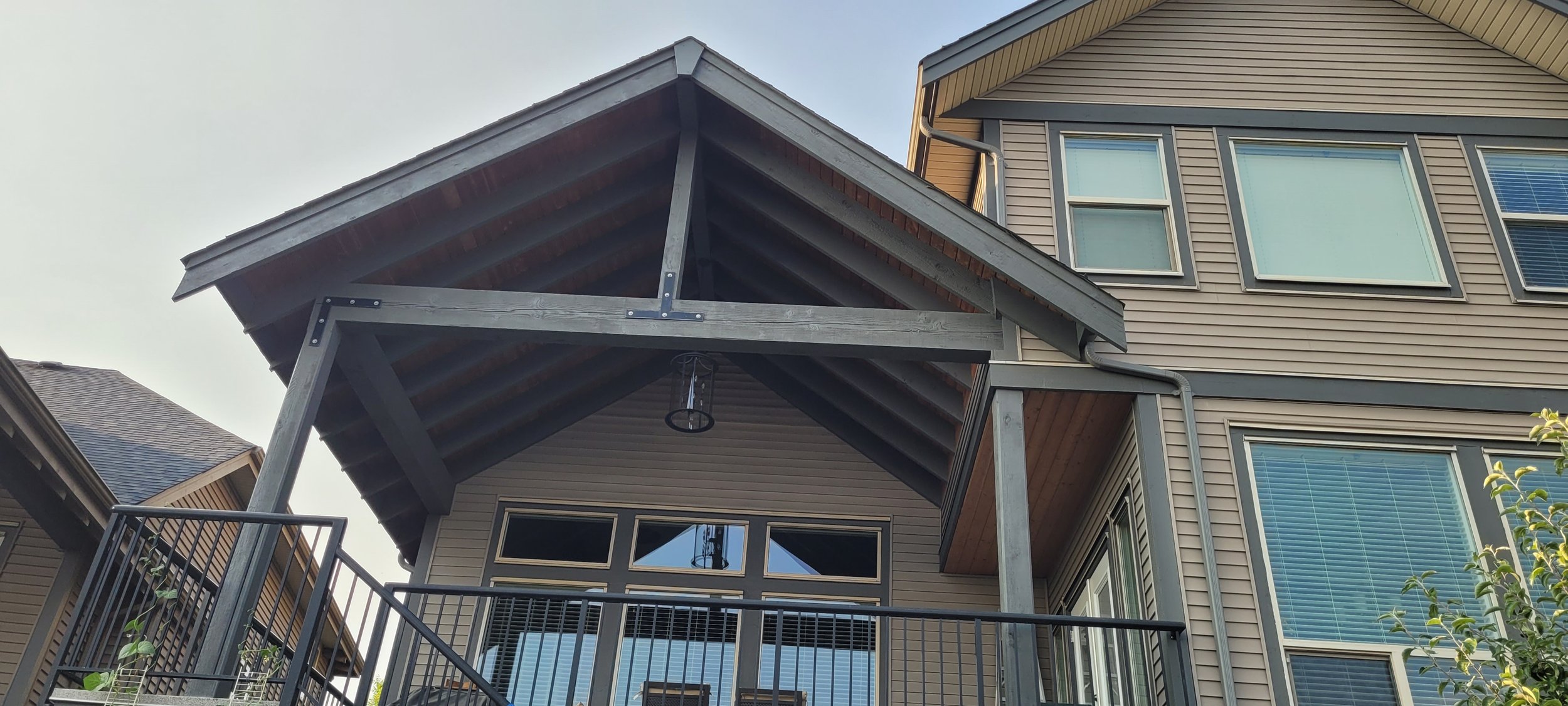 Back view of a modern house with a covered patio and balcony, featuring a gabled roof, multiple windows, and a metal railing.