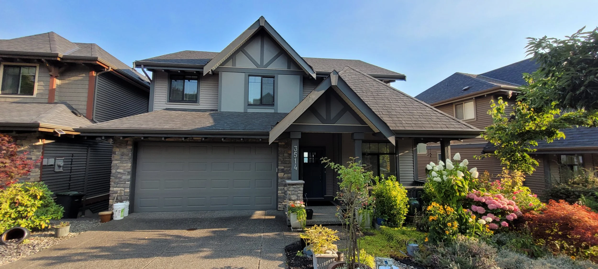 A modern two-story house with gray siding and stone accents, with a landscaped front yard featuring colorful flowers and shrubs, and neighboring houses visible on either side.