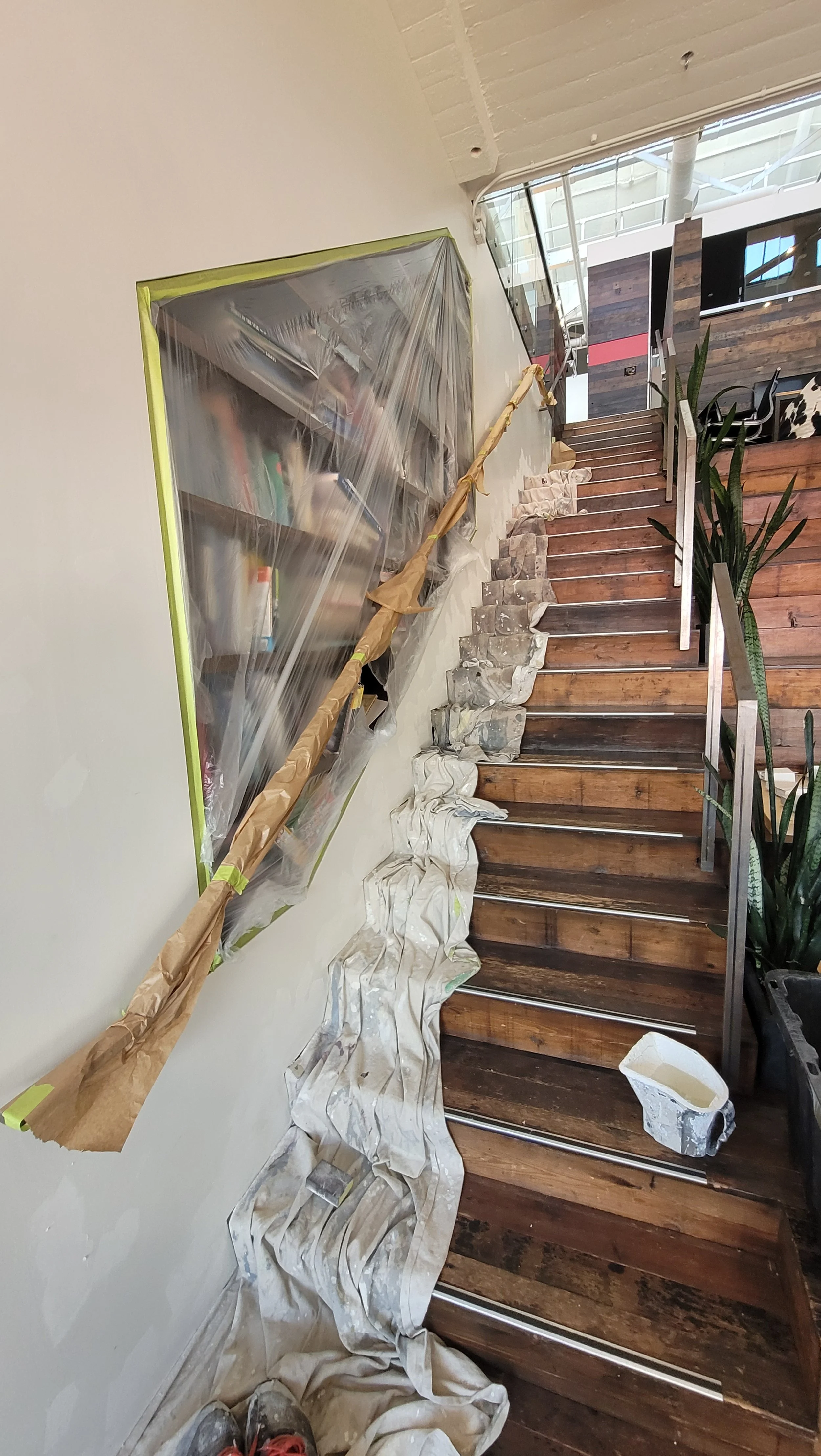 Indoor staircase undergoing renovation with protective covering, painting supplies, and tools placed on the floor.