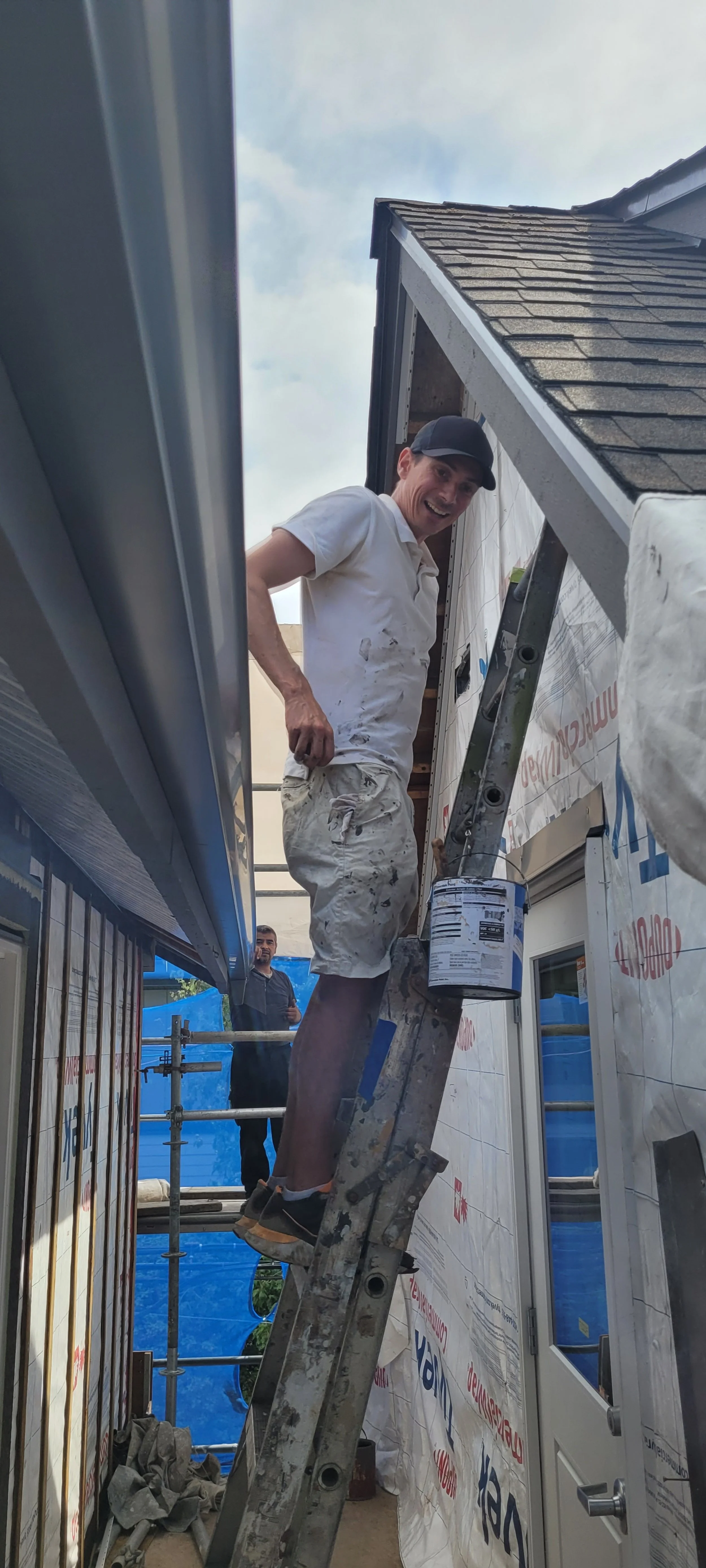 A man in white painter's clothes smiling while standing on a ladder working on the exterior of a house under construction or renovation. Behind him, another person stands on scaffolding.