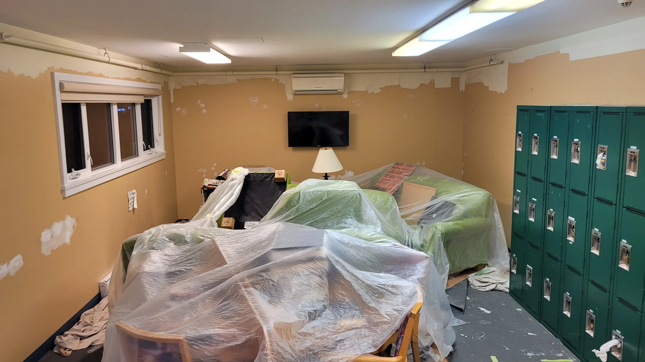 Room with partially painted tan walls, a window on the left, a television on the back wall, green lockers on the right, and furniture covered with plastic sheets.