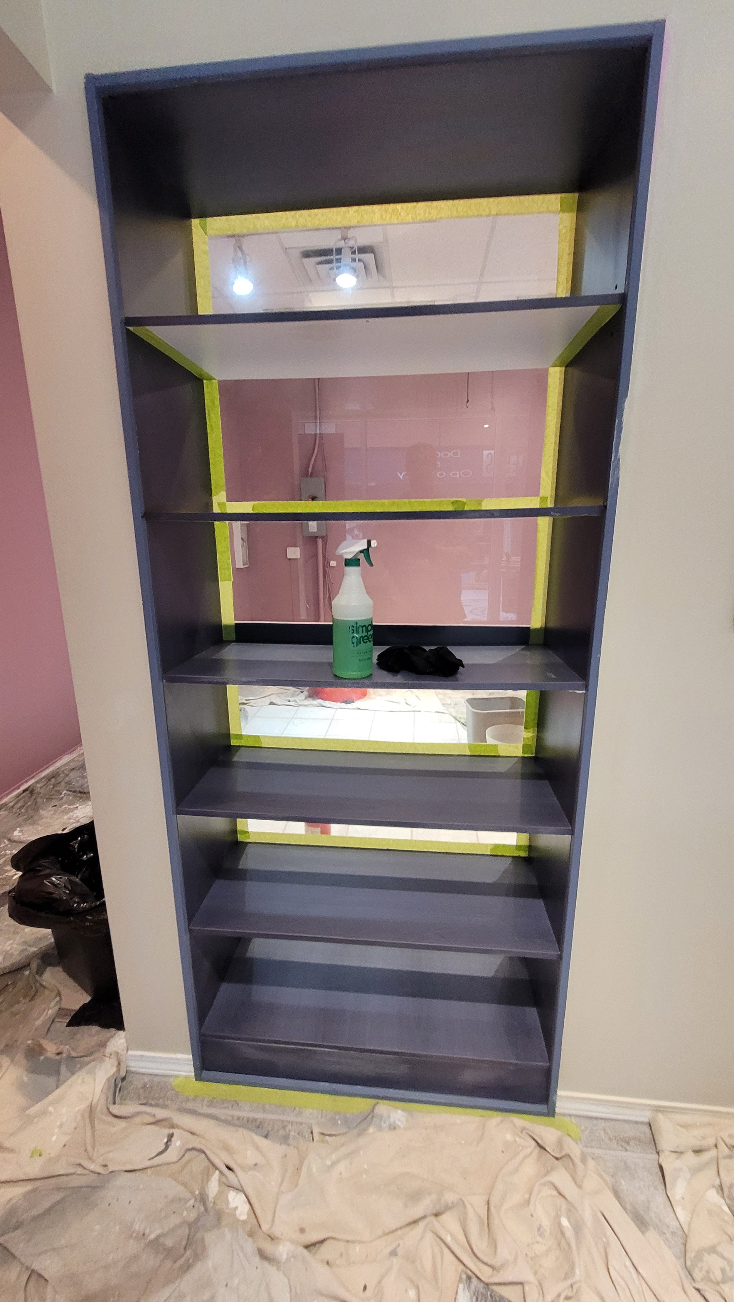 An empty bookshelf with six shelves, a mirror behind it with yellow tape around the edges, a spray bottle, and black gloves on one of the shelves. The wall and ceiling are visible in the background, and the floor is covered with protective sheets and