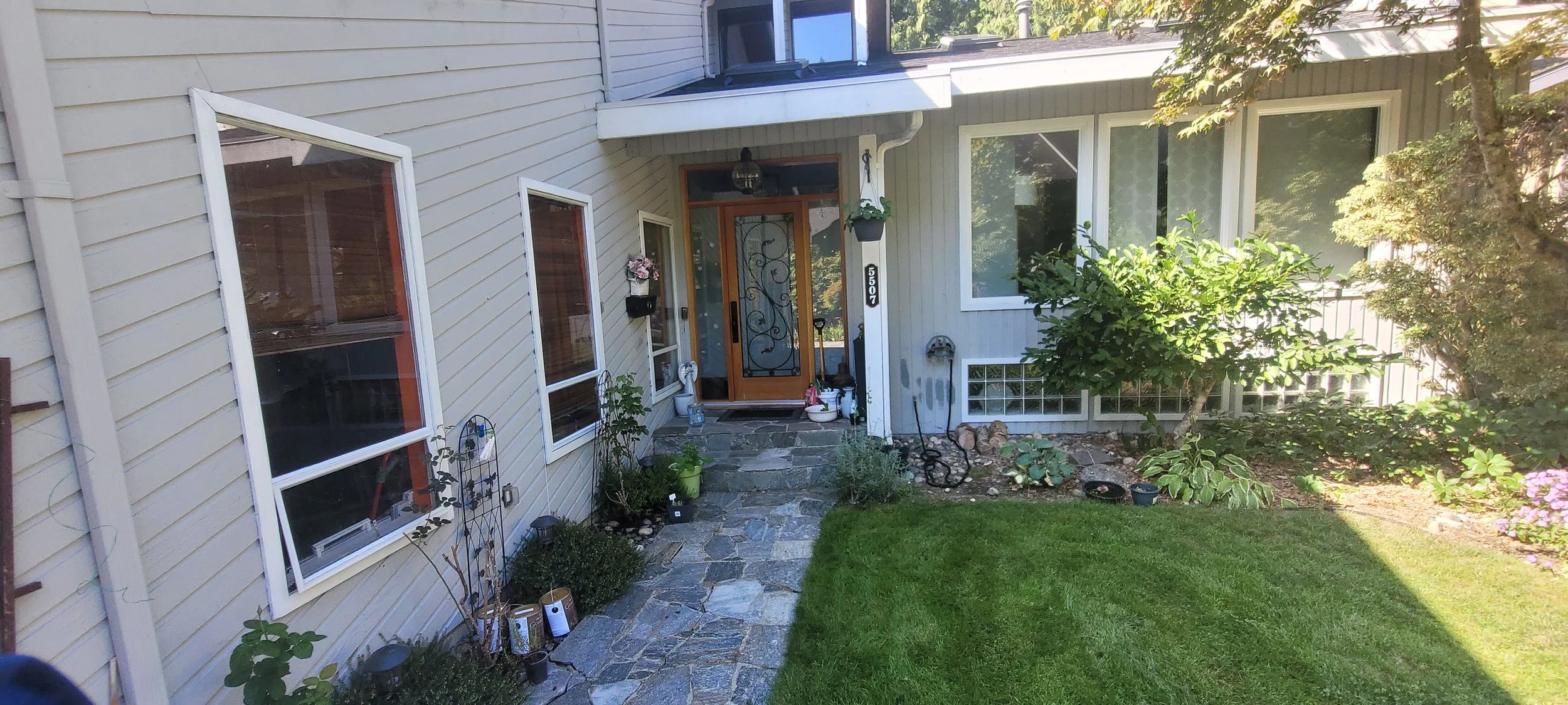 A view of a house's front yard with a stone pathway leading to the front door. The house has beige siding, large windows, and a small garden with plants and trees. The front door is wooden with decorative ironwork, and there are potted plants and gar