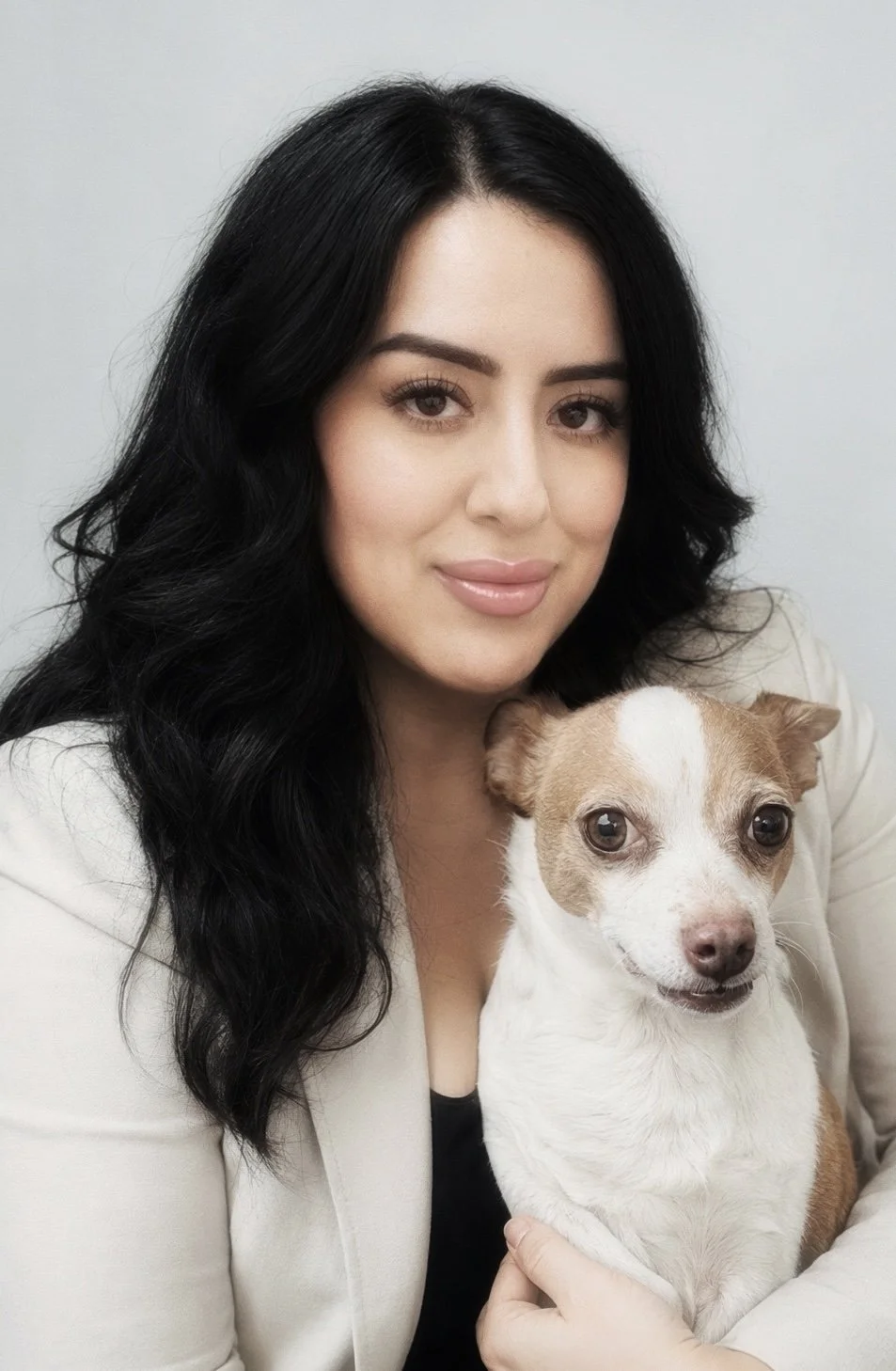 A woman with long black hair holding a small tan and white dog, both looking at the camera against a light gray background.
