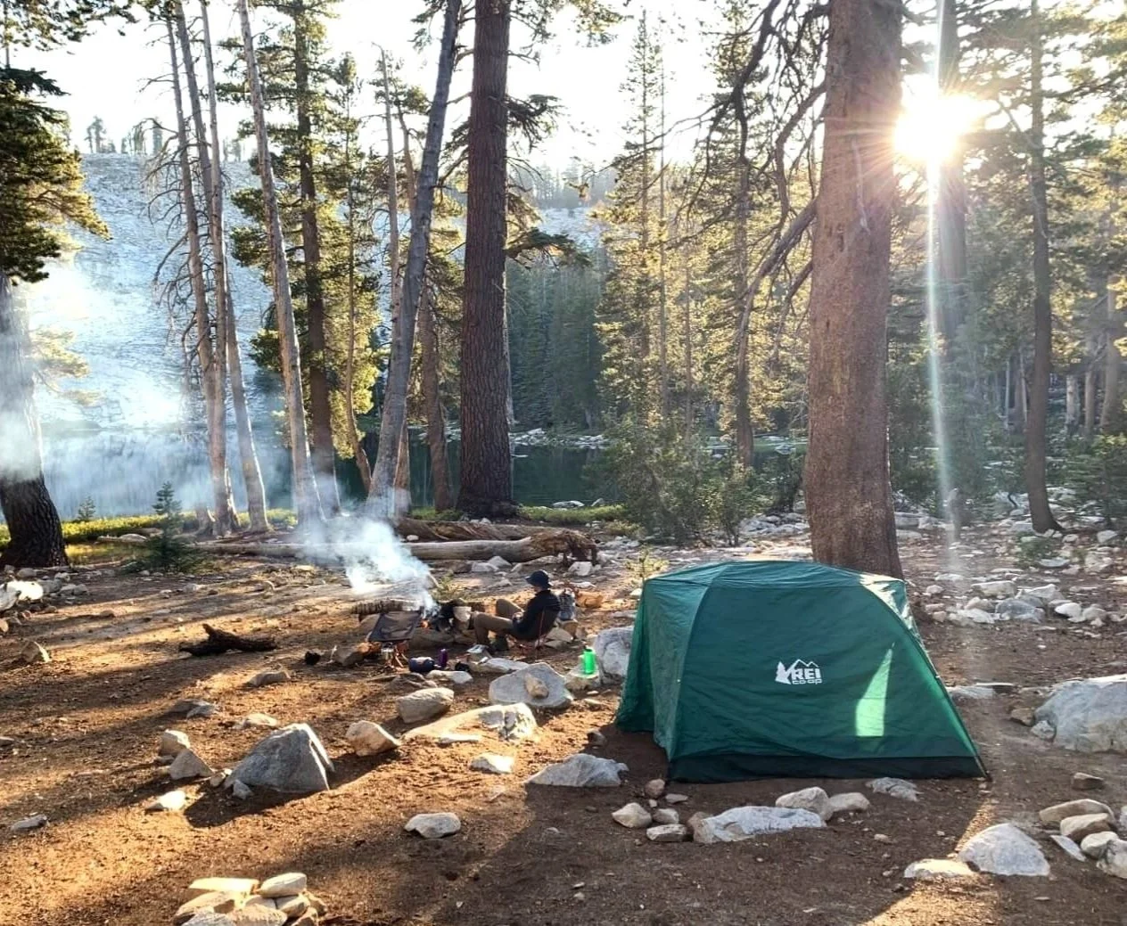 A camping scene in a forest with tall trees, a green tent, and a campfire with smoke, near a lake with the sunlight shining through the trees.