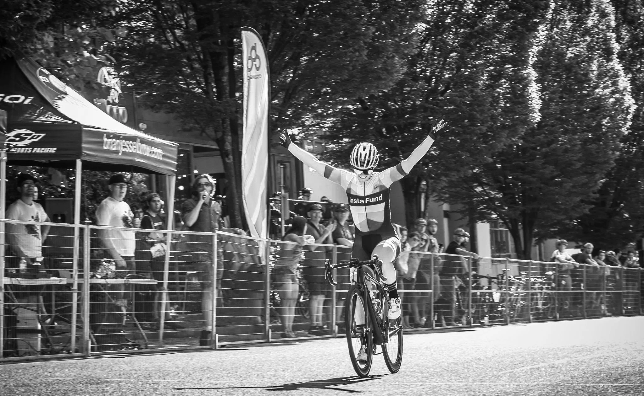 A cyclist crossing the finish line with arms raised in victory, on a city street lined with spectators and trees.