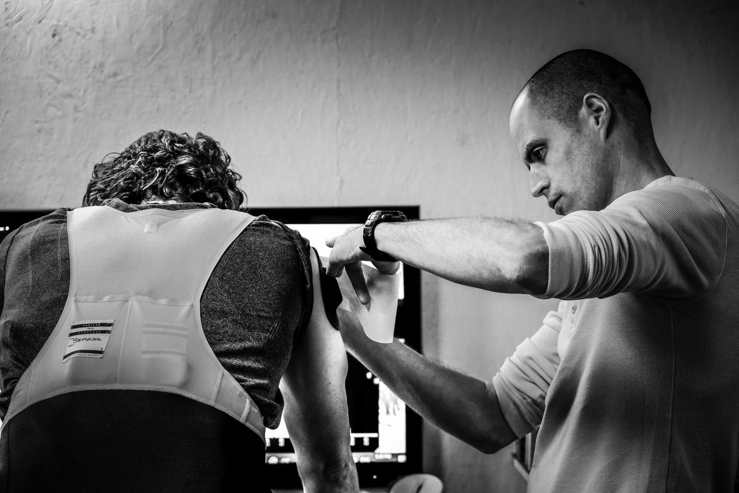 A man with a shaved head providing An injection to a firefighter with curly hair, who is wearing a vest, in an indoor setting with a computer monitor in the background.