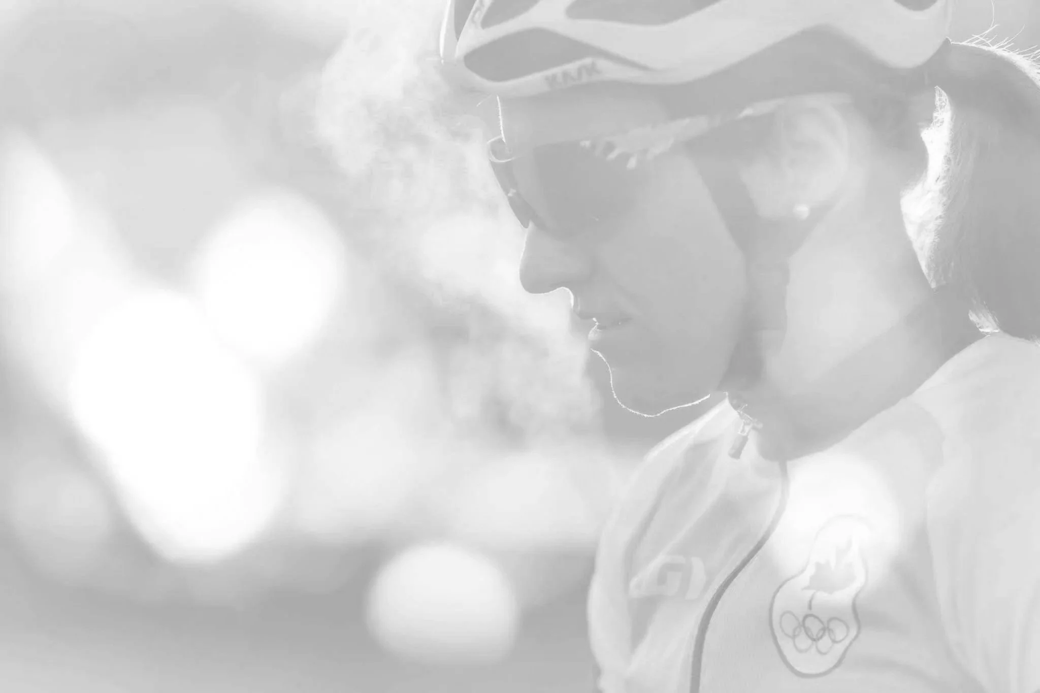 A female athlete wearing glasses and a jacket with the California Olympic logo, looking down and appearing focused, with a blurred background of lights.