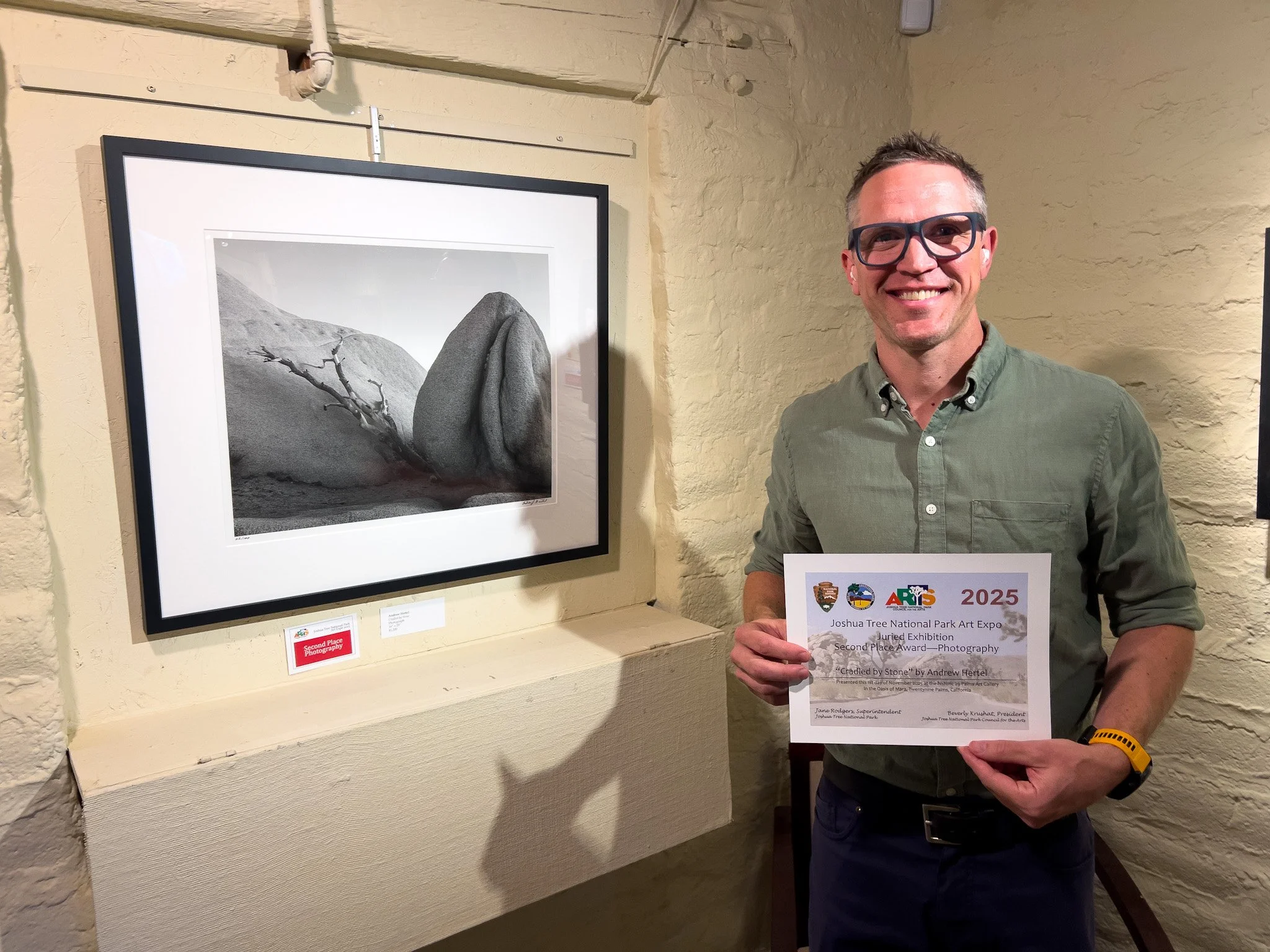 Man, Andrew Hertel, standing next to photograph, holding an award received for the photograph at an art gallery.