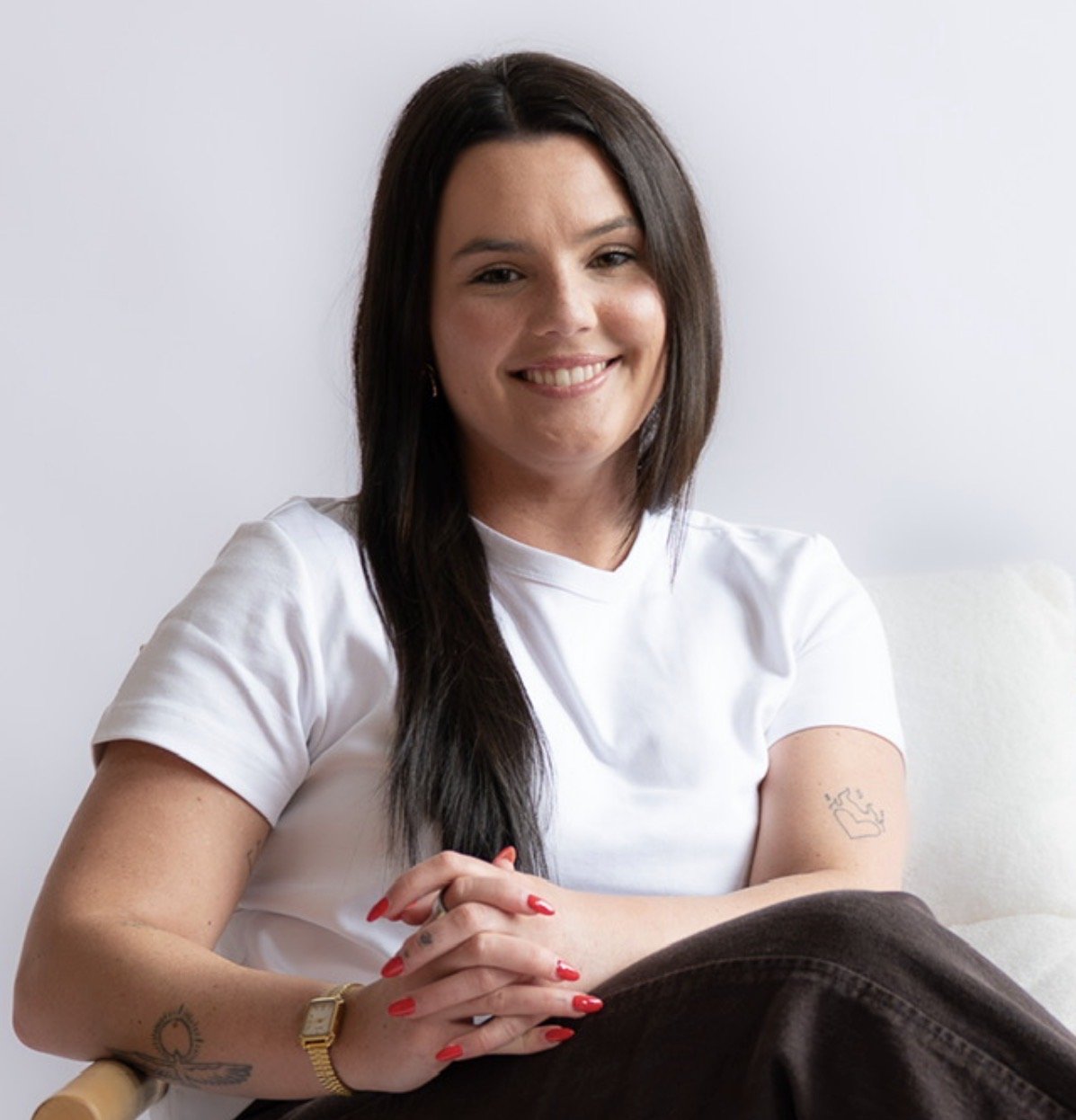 A woman with long dark hair, smiling, wearing a white top, sitting on a couch in a cozy living room with hardwood floors, a lamp, and a cat resting on a blanket behind her.