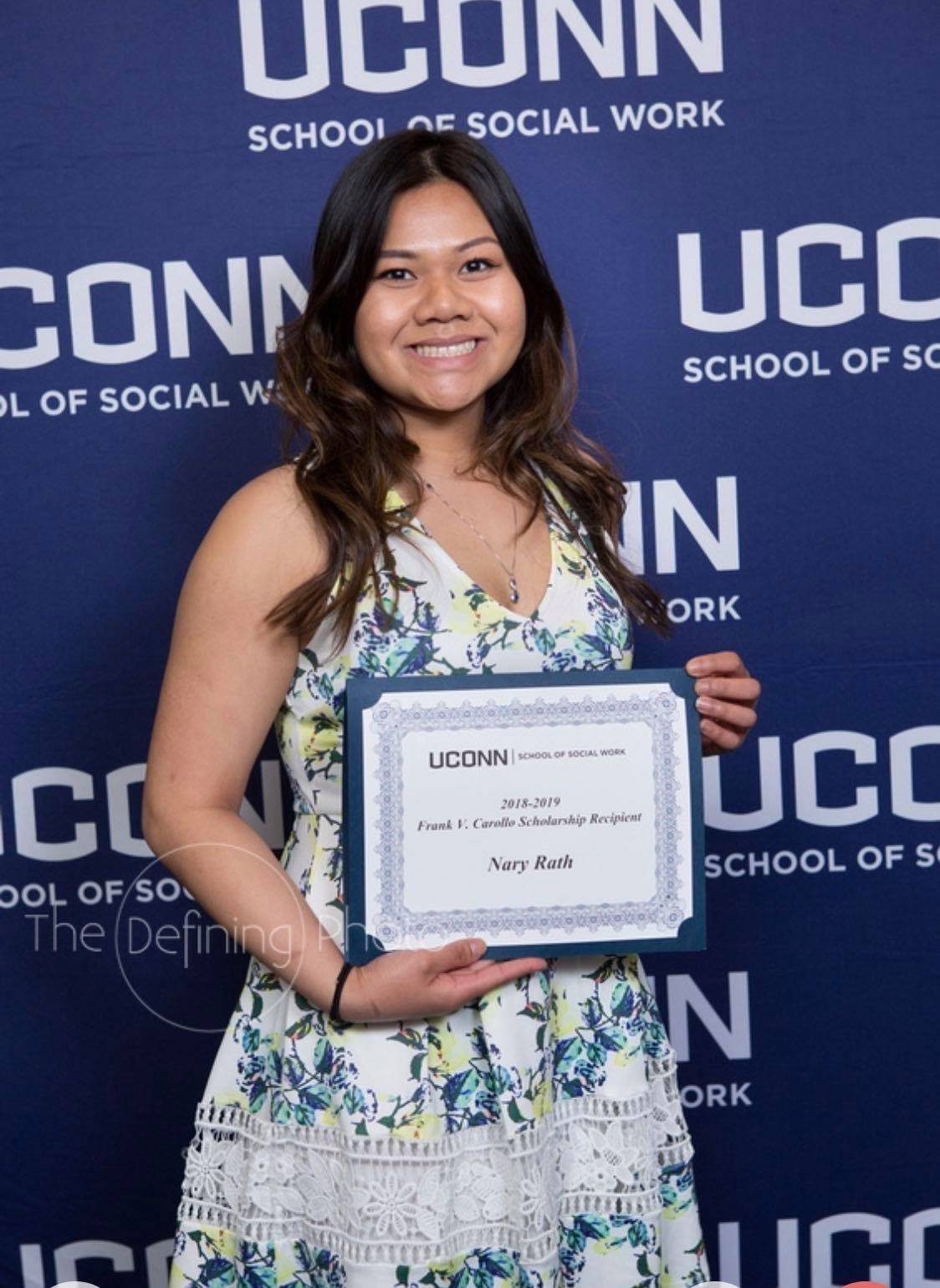 A young woman smiling and holding a certificate in front of a blue backdrop with 'UCONN School of Social Work' logos. She has long wavy brown hair and is wearing a floral dress.