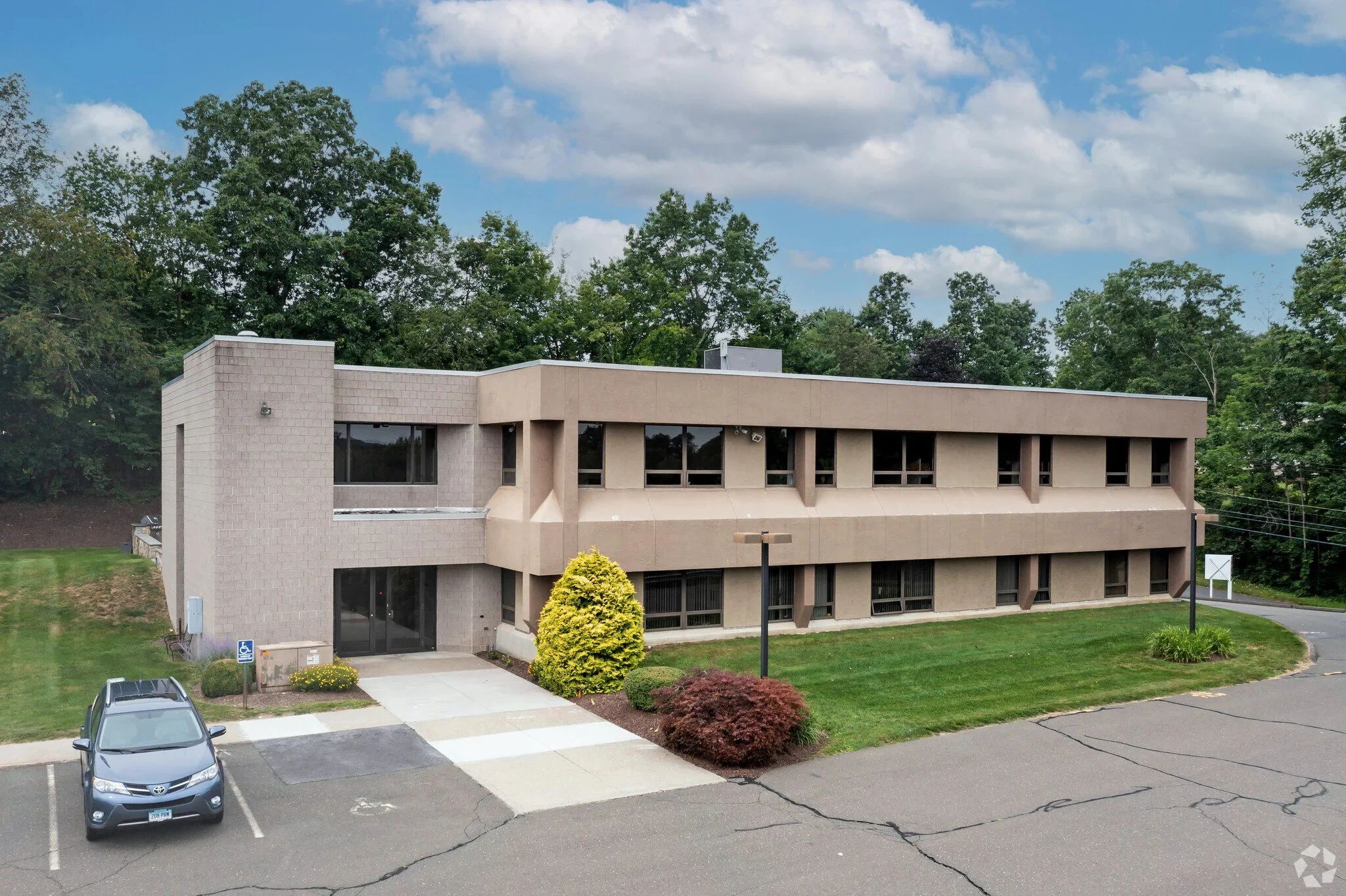 A beige multi-story office building with large glass windows and a flat roof, surrounded by a parking lot, green lawns, and landscaped bushes and trees. Cloudy sky in the background.