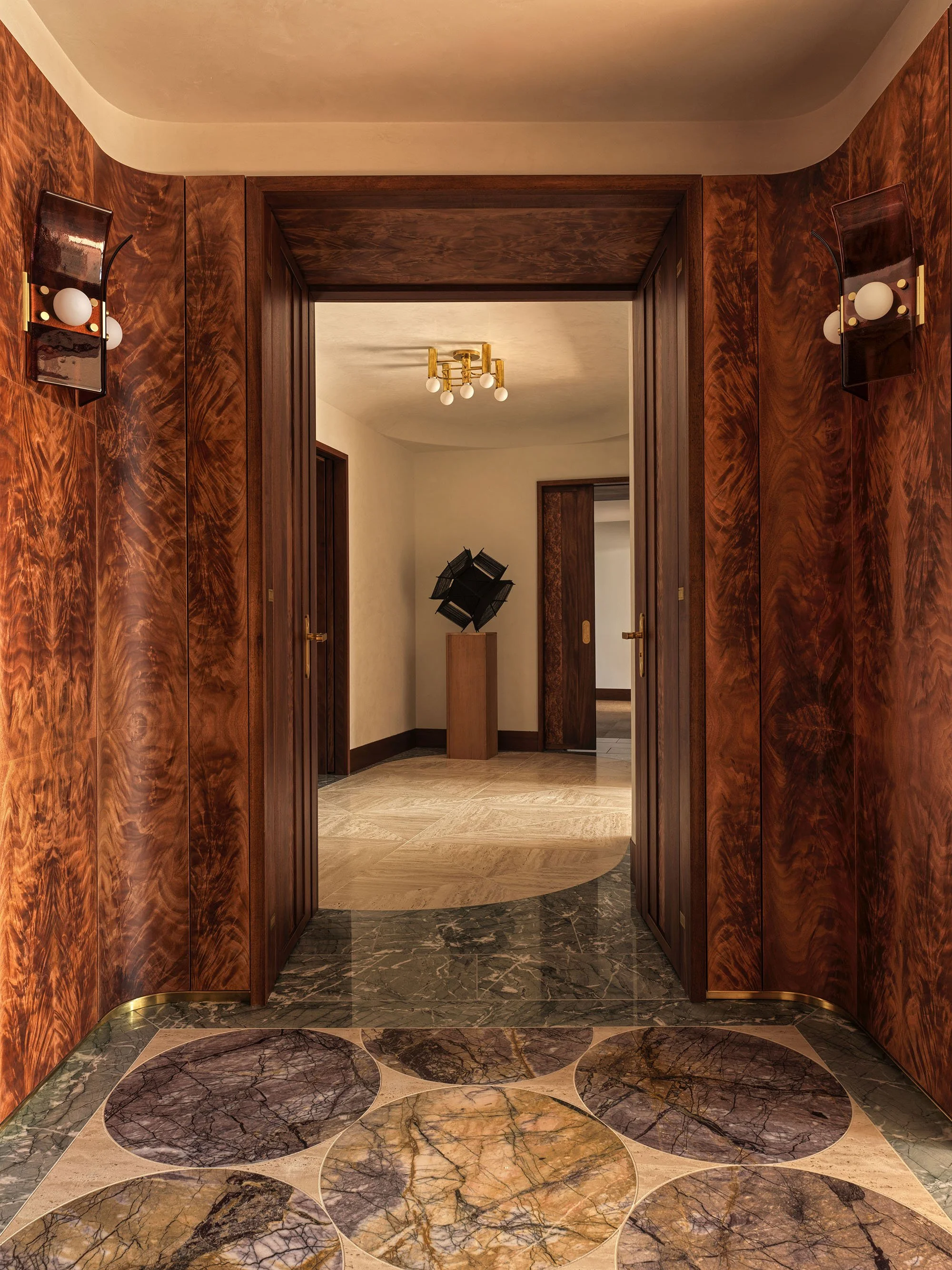 A vestibule featuring curved okoume paneling and a floor of lilac marble roundels set in travertine leading to a gallery with an abstract sculpture