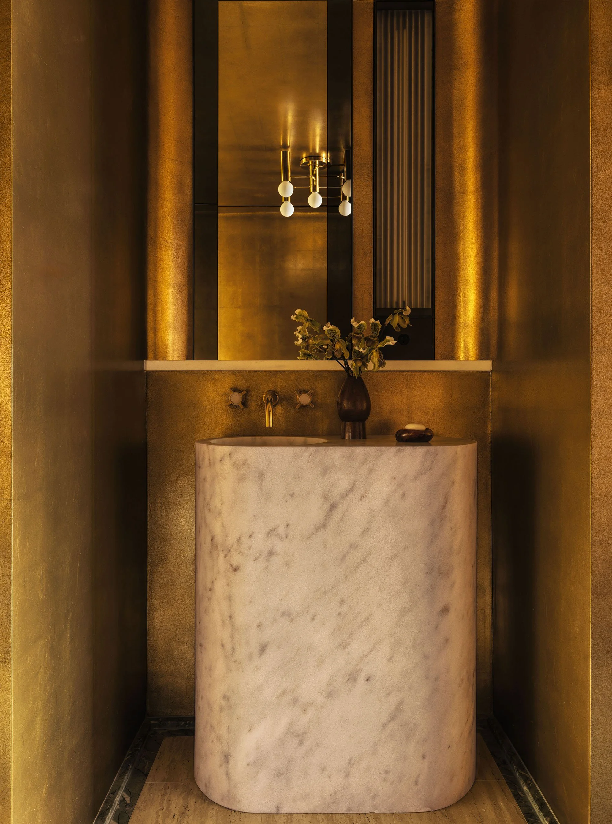 A powder room with curved gold-leaf walls and ceilings, a cylindrical marble sink, and a vintage brass ceiling fixture