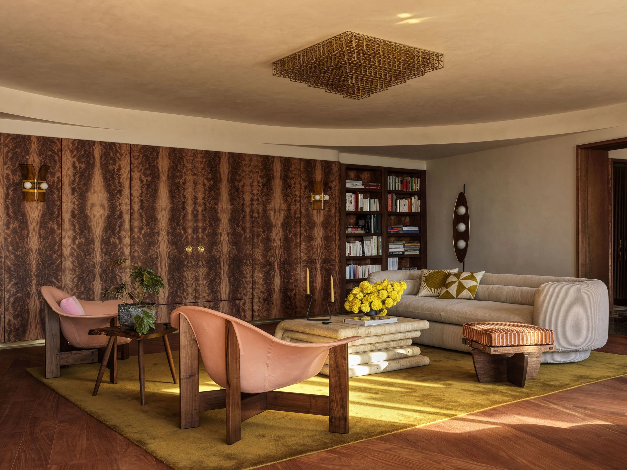 A sunlit living room featuring a bookmatched okoume wall, an ecru corduroy sofa, and blush pink leather armchairs under a gridded brass light fixture