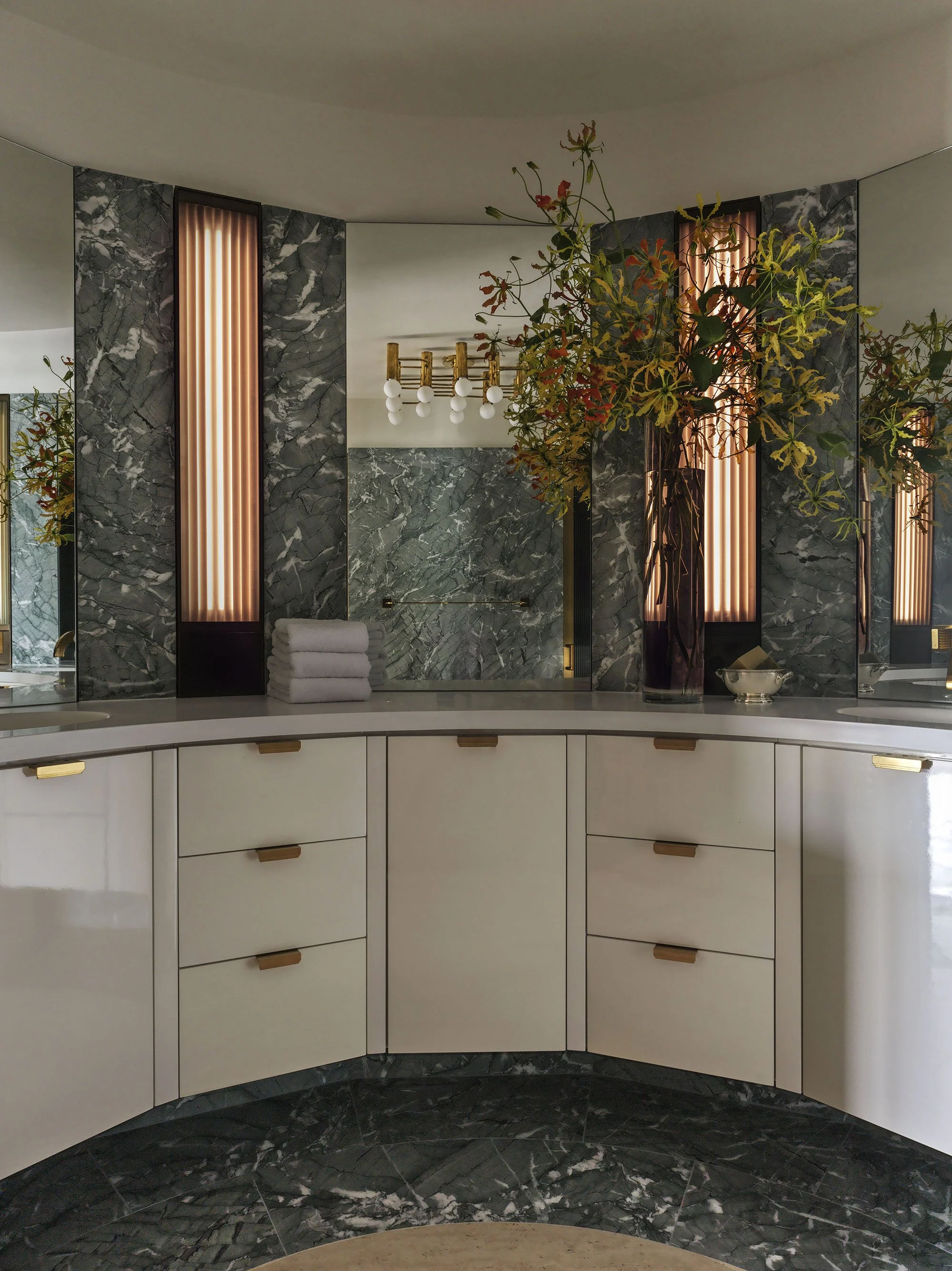 A curved floating vanity in the Watergate apartment designed by Nicholas Potts featuring a floating white lacquer curved vanity, under curved green marble walls and mirrors featuring bronzed sconces, a reflection of vintage brass light fixture
