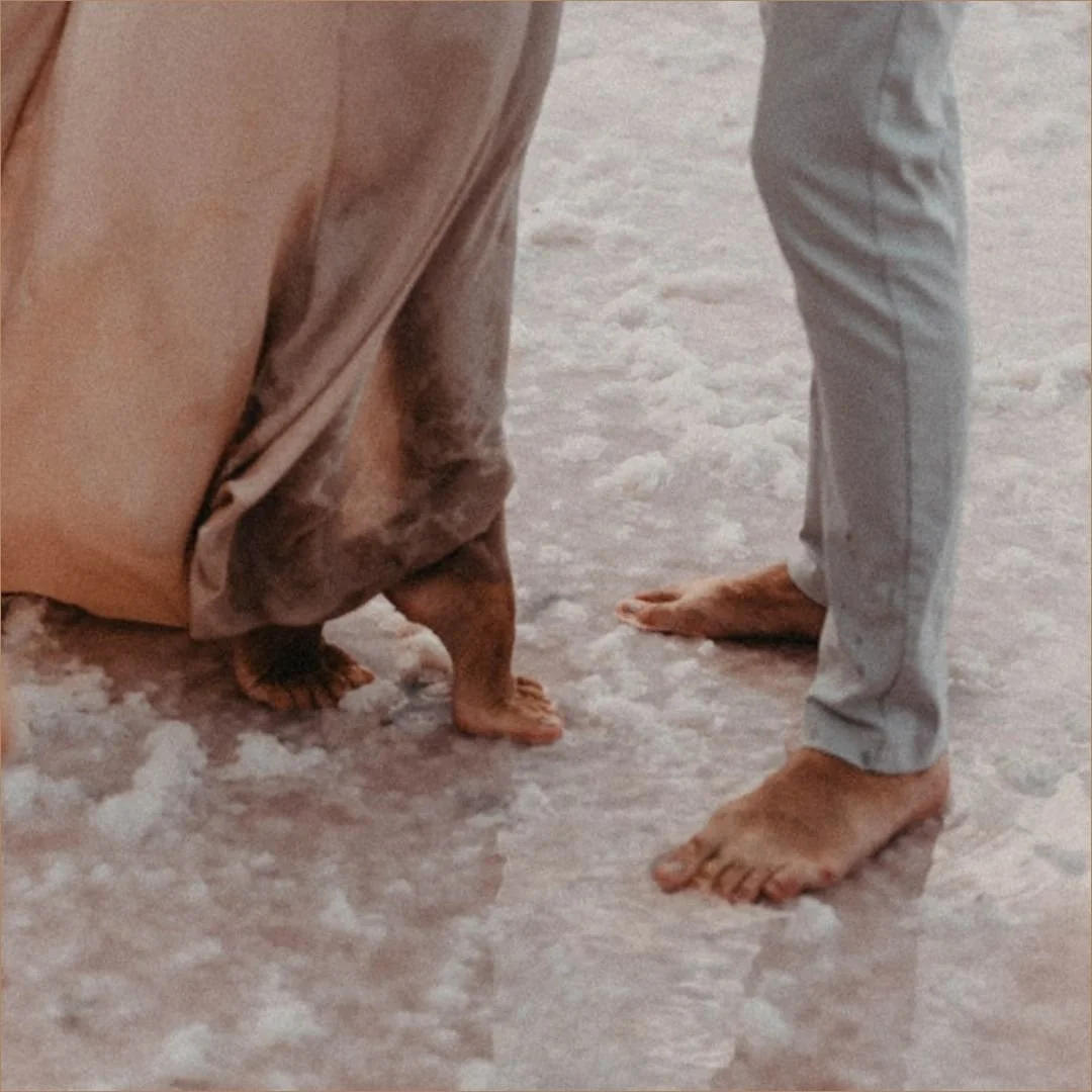 A man and woman standing barefoot in shallow coastal water.