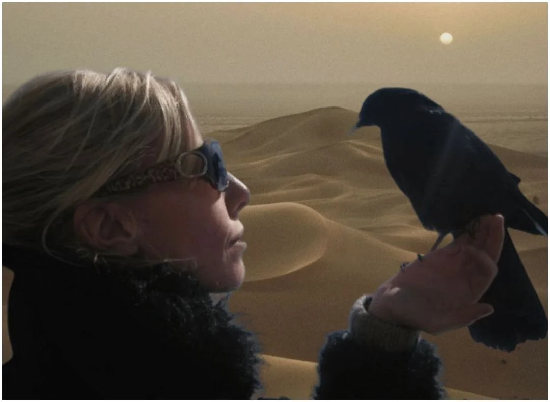 A portrait of Elisabeth Paul outdoors in nature, looking at a black bird perched on her hand against a background of sand dunes.