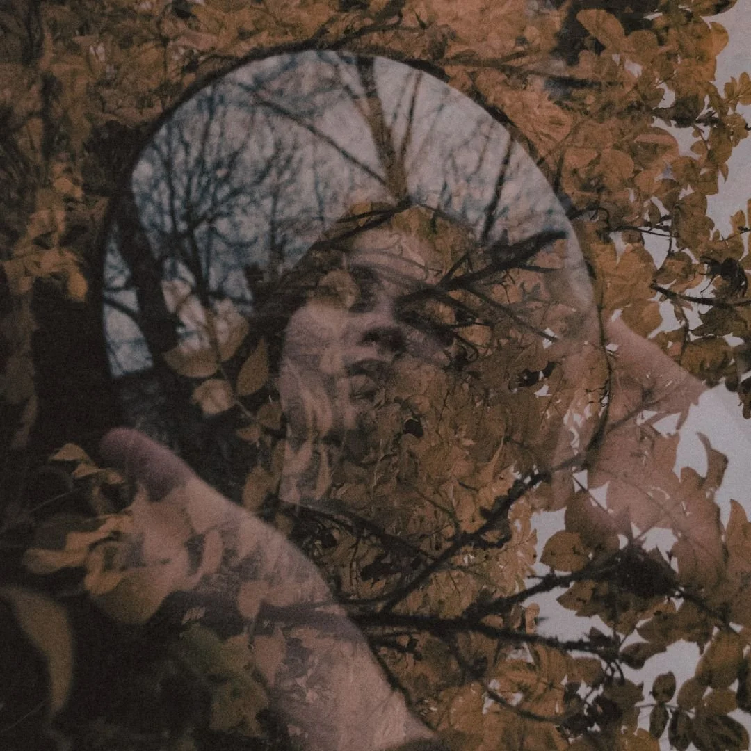 A woman looking into a handheld mirror surrounded by brown leaves, with a subtle reflection of her face visible.