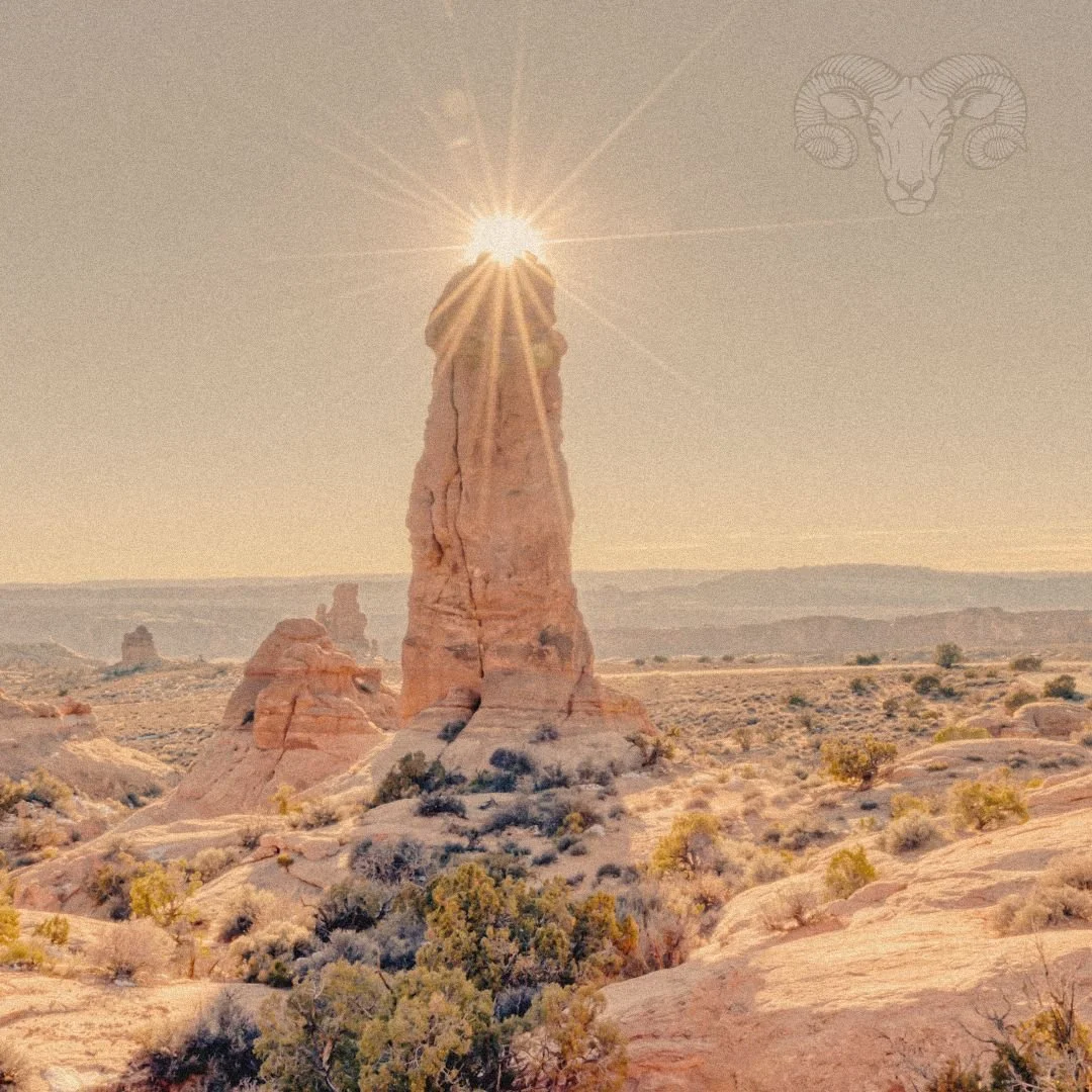A pillar rock formation with the sun beaming behind the top, surrounded by red, rocky terrain.