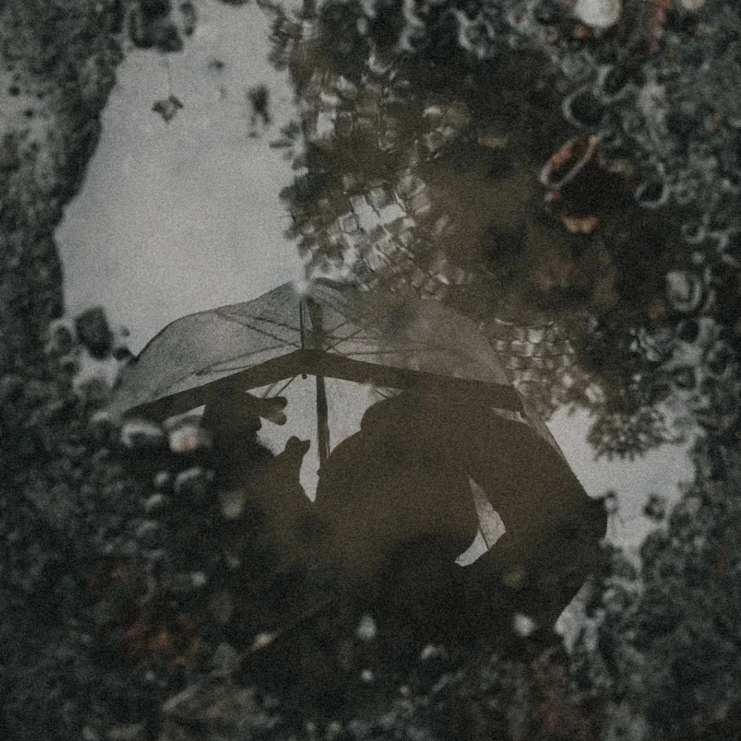 A reflection in a water puddle on wet ground, showing the dark silhouette of a person holding an open umbrella.