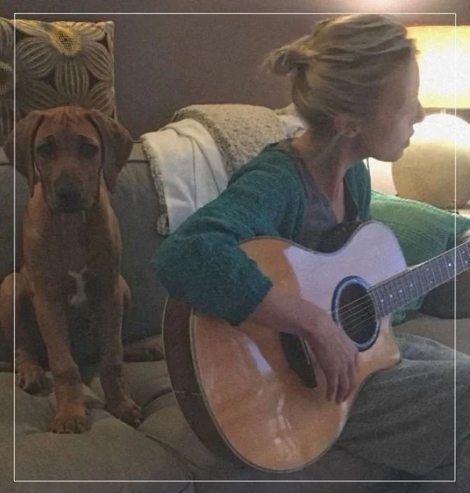 A casual portrait of Elisabeth Paul sitting with her dog while playing an acoustic guitar.