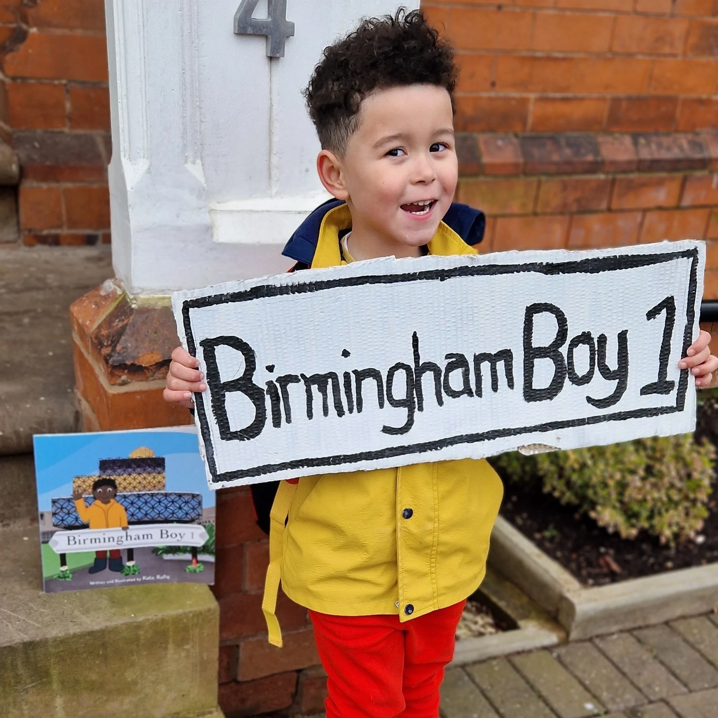 A young boy with curly hair wearing a yellow jacket and red pants is holding a sign that reads 'Birmingham Boy 1'. He is smiling and standing outside near a brick wall, next to a small illustrated sign of himself with the same text.